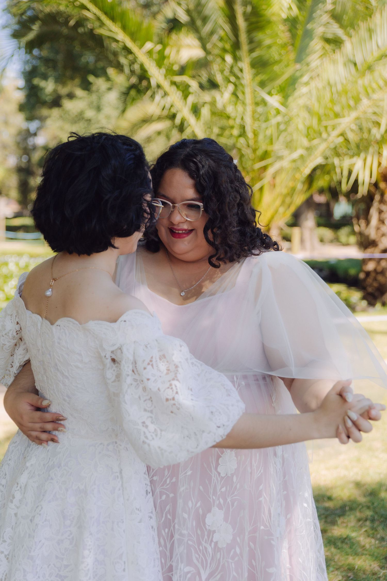 Two brides in white and blush lace wedding gowns share a joyful embrace in a garden.