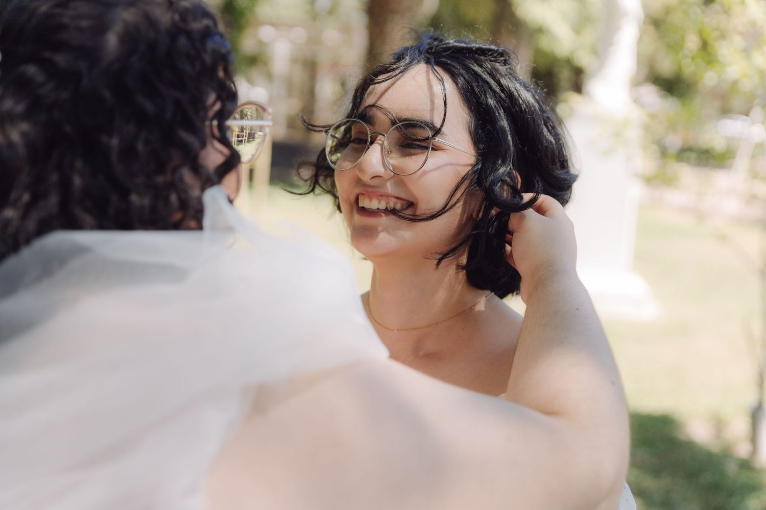 Close-up of two brides sharing an intimate moment outdoors, one wearing glasses and smiling warmly.