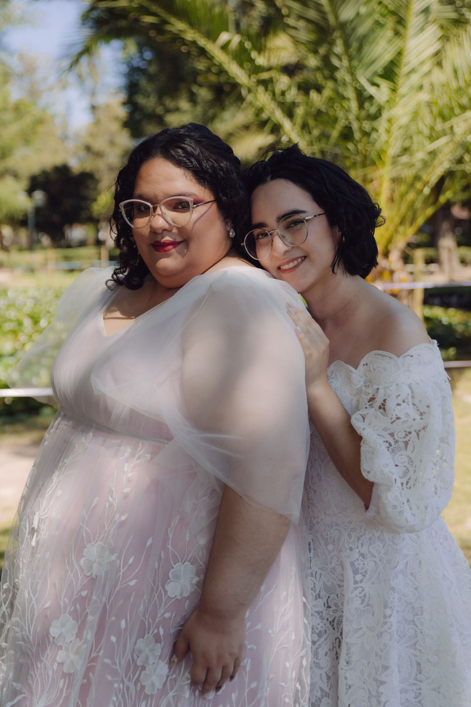 Two brides in white and blush lace wedding dresses pose back-to-back smiling in a sunny garden.
