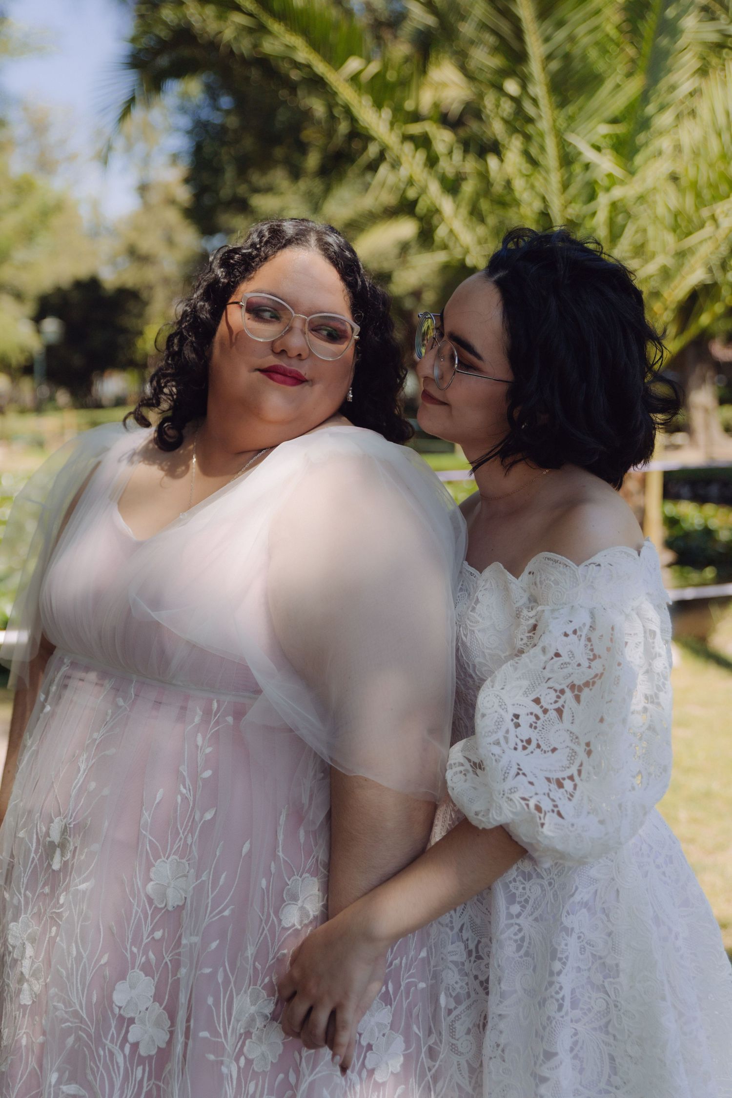 Two brides in white wedding dresses embrace outdoors, one in lace off-shoulder gown, other in floral tulle dress, both wearing glasses.