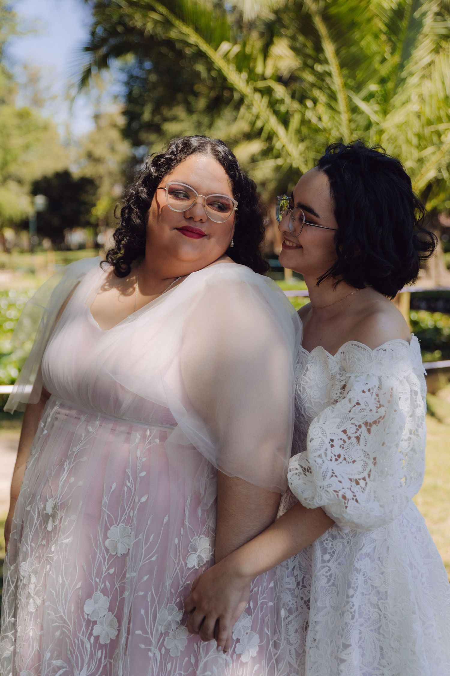 Two brides sharing an intimate moment in garden setting, wearing elegant white wedding dresses and glasses on sunny day.
