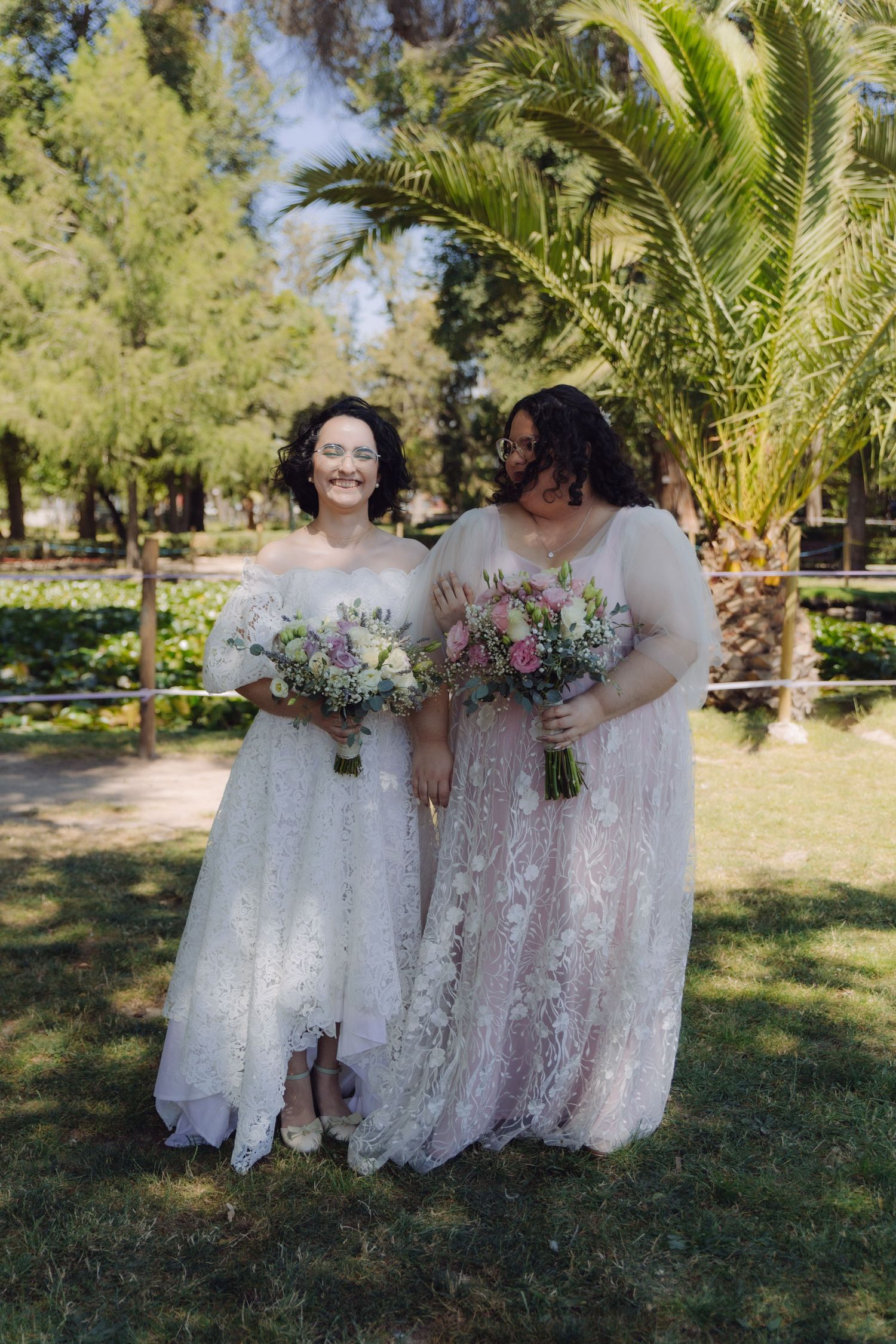 Two brides standing together in garden holding floral bouquets, wearing white lace and pink embroidered wedding gowns outdoors.