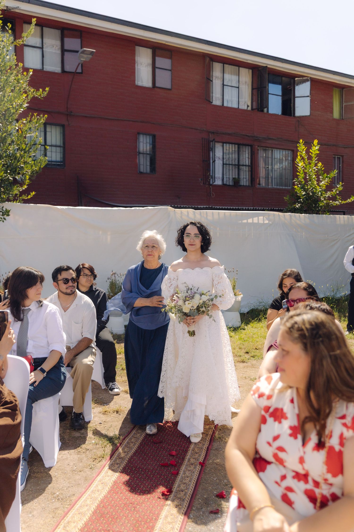 Bride in white off-shoulder lace gown walks down outdoor aisle with older woman, carrying white floral bouquet at wedding.