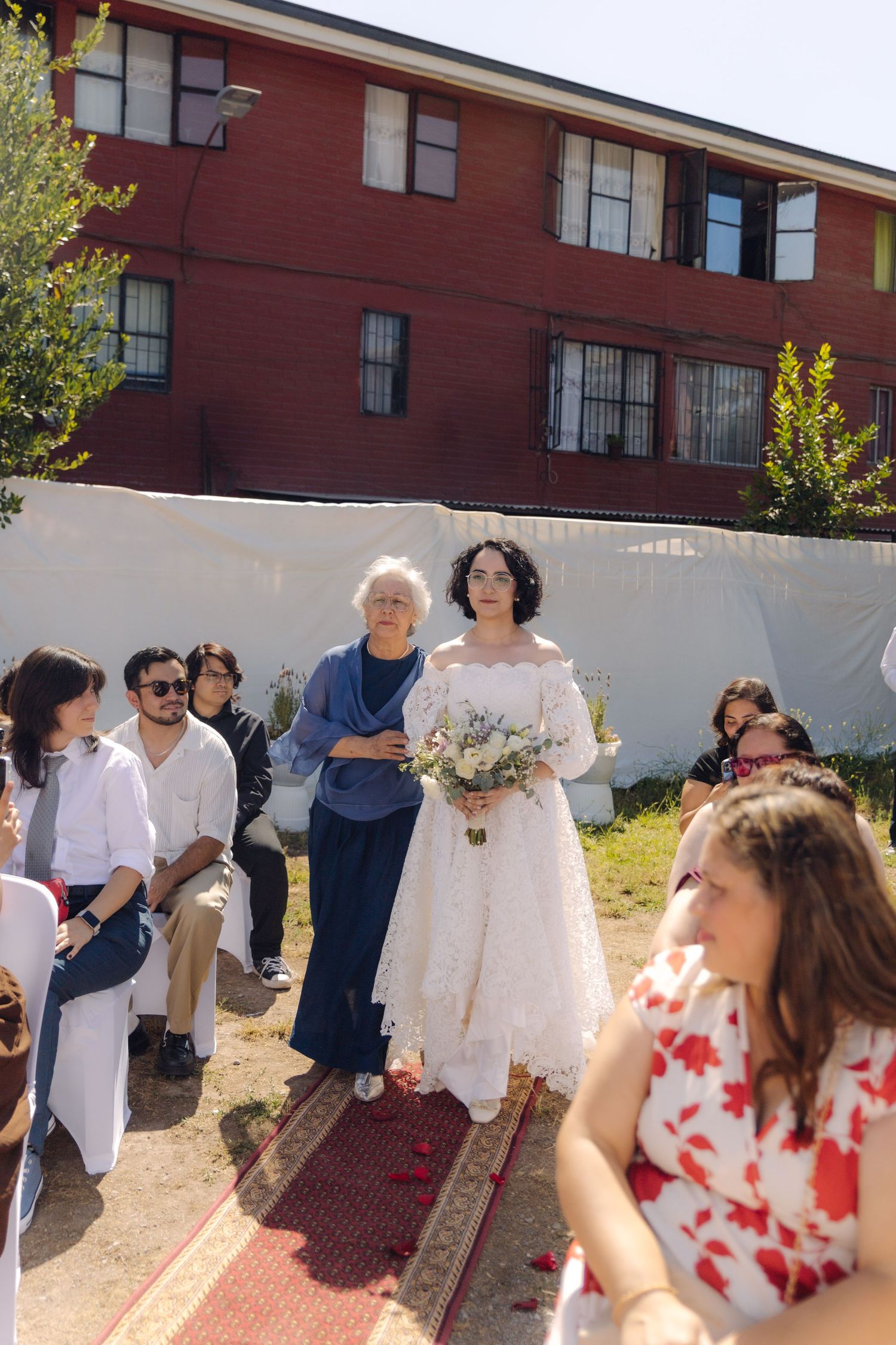 Bride in white off-shoulder lace gown and elderly companion walk down outdoor aisle at sunny wedding ceremony with guests.