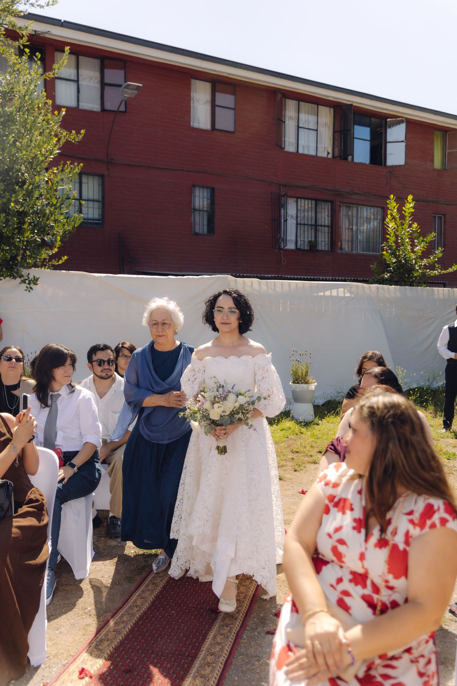 Bride in white lace off-shoulder wedding dress walks aisle with elderly companion, holding bouquet at outdoor ceremony.