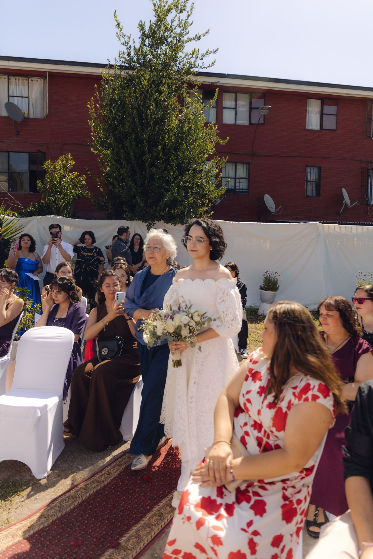 Bride in white lace gown walks down red carpet aisle with older woman at sunny outdoor wedding ceremony with guests.