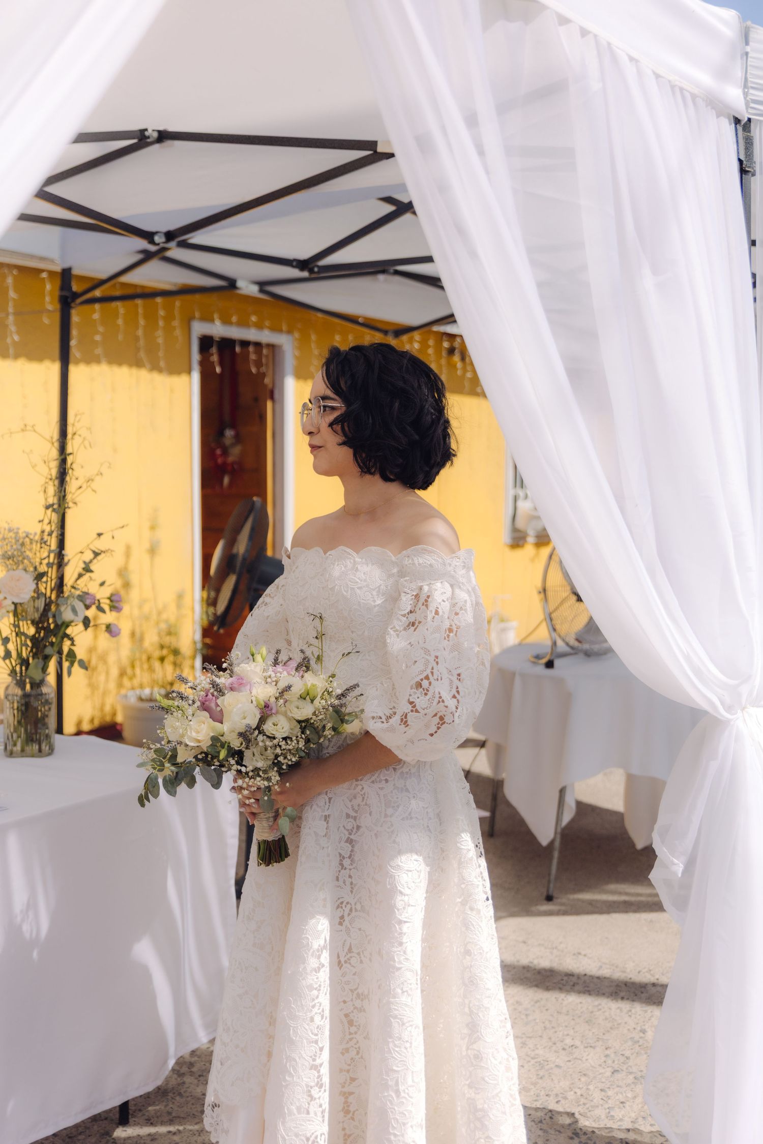 Bride in white off-shoulder lace wedding dress holds floral bouquet while standing under white draped canopy at ceremony.