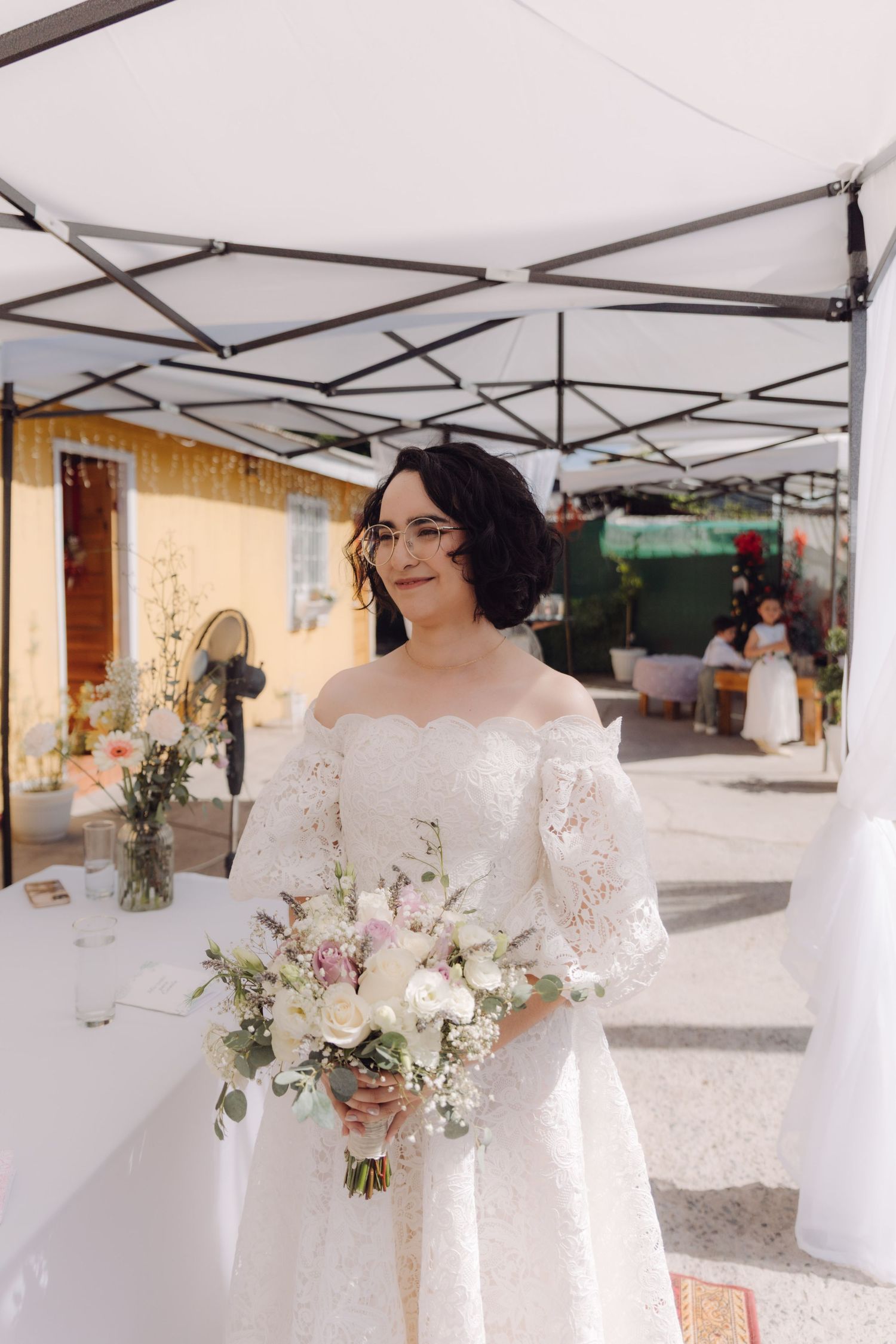 Bride in white off-shoulder lace wedding dress smiles while holding pastel floral bouquet under canopy at outdoor ceremony.