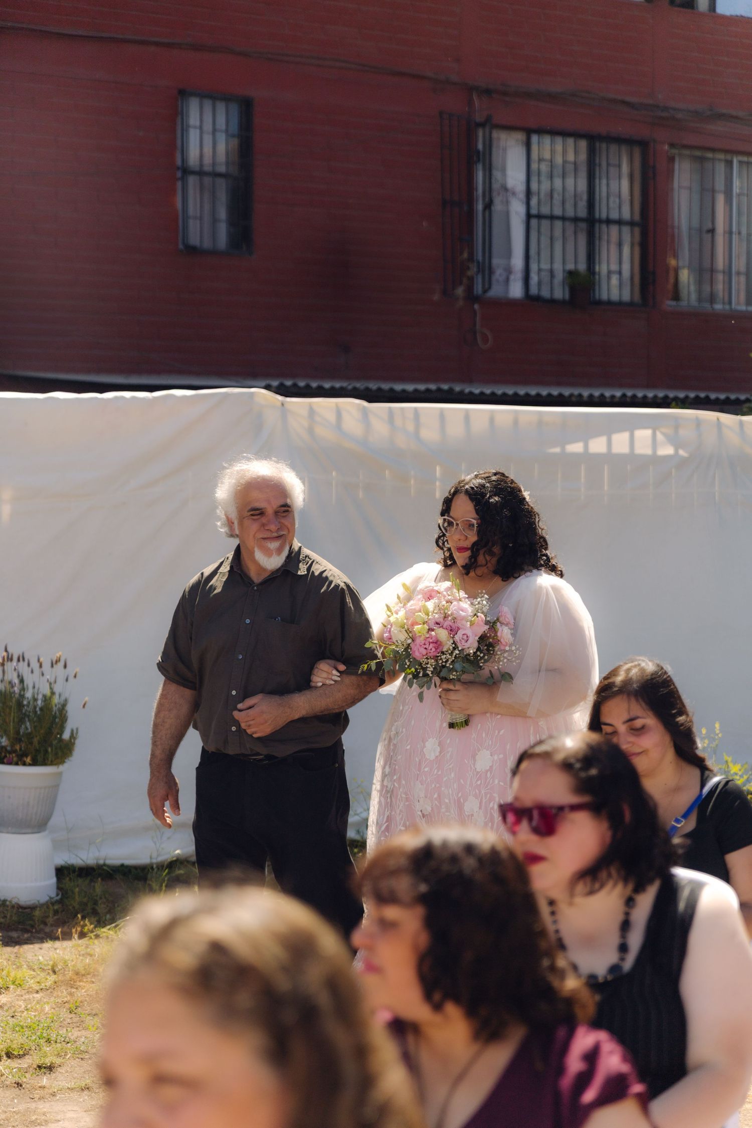 Plus-size bride in pink floral tulle wedding gown walks down aisle with older gentleman at outdoor wedding ceremony.