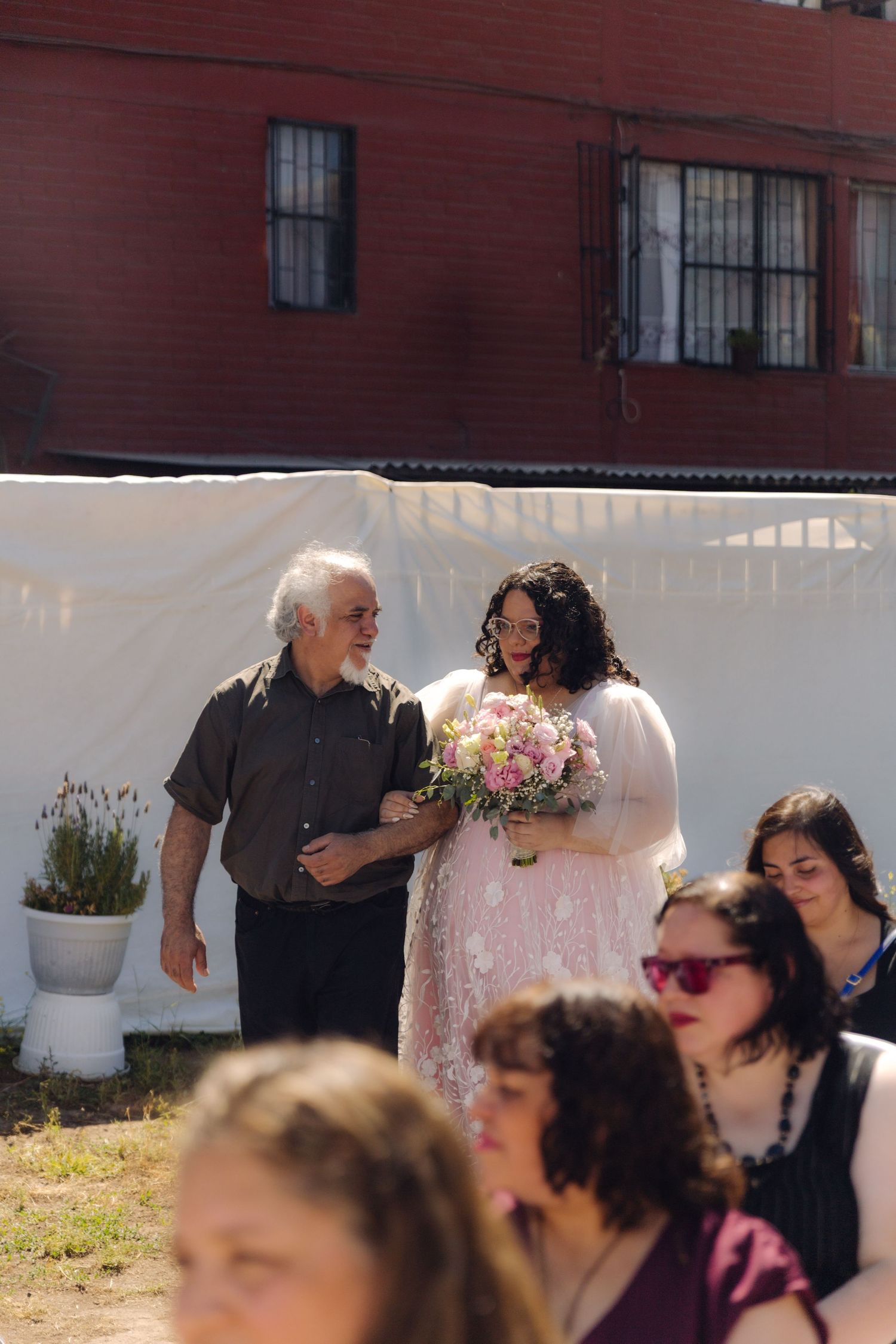 Bride in pink lace gown walks down aisle with elderly escort at outdoor wedding ceremony.