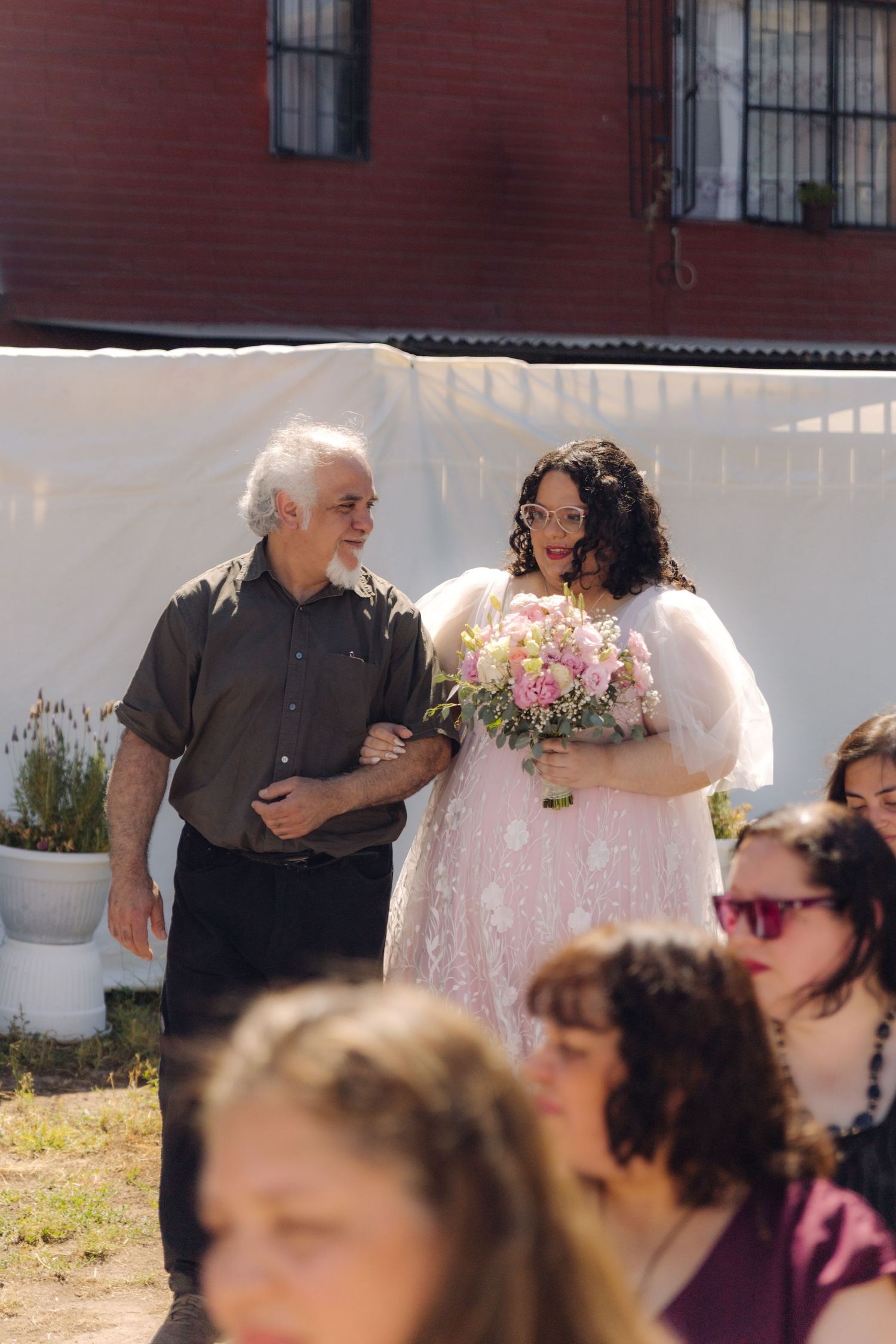 Father escorts bride in pink floral gown holding bouquet at sunny outdoor wedding ceremony.