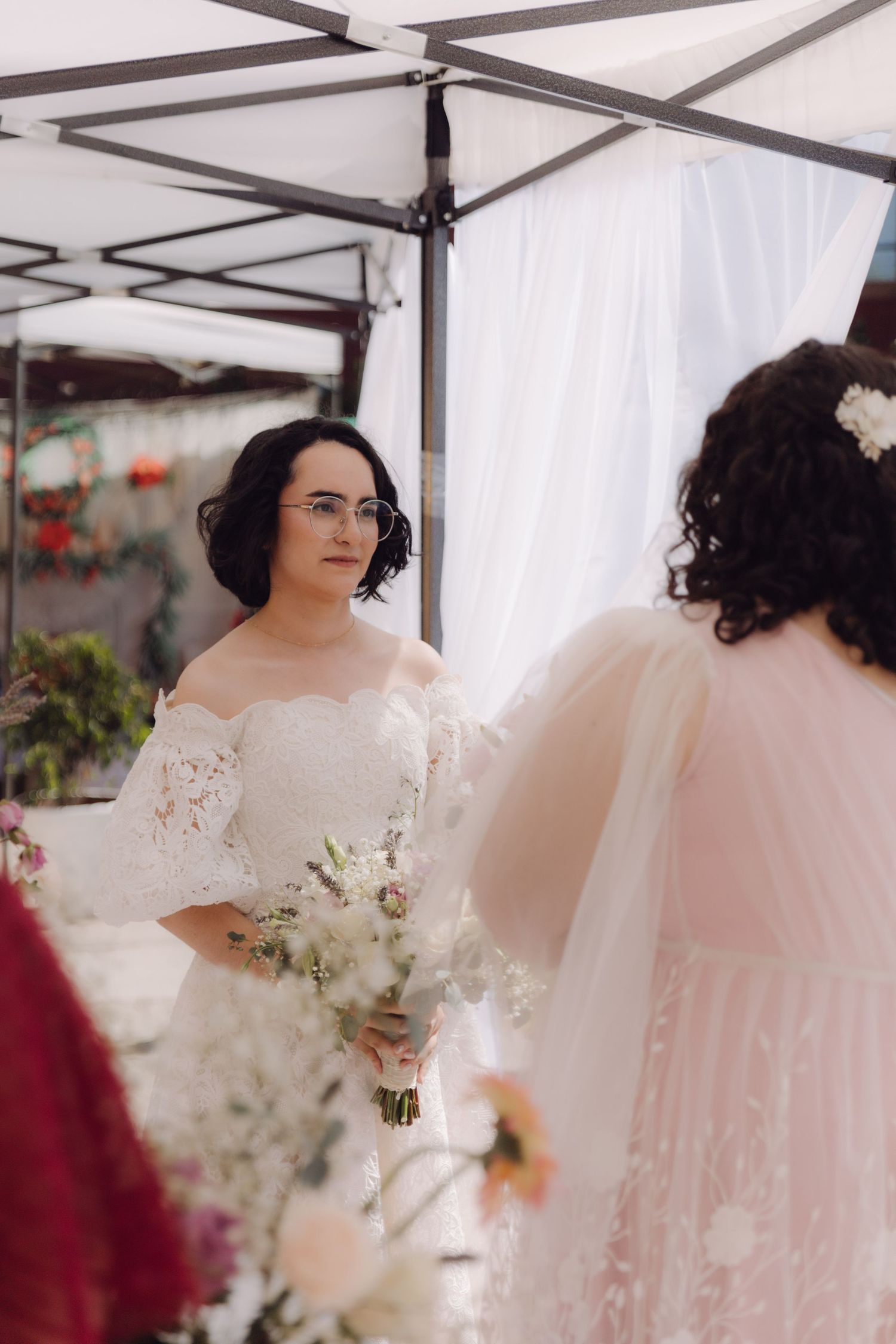 Bride in white off-shoulder lace gown holds bouquet facing partner at outdoor wedding altar.