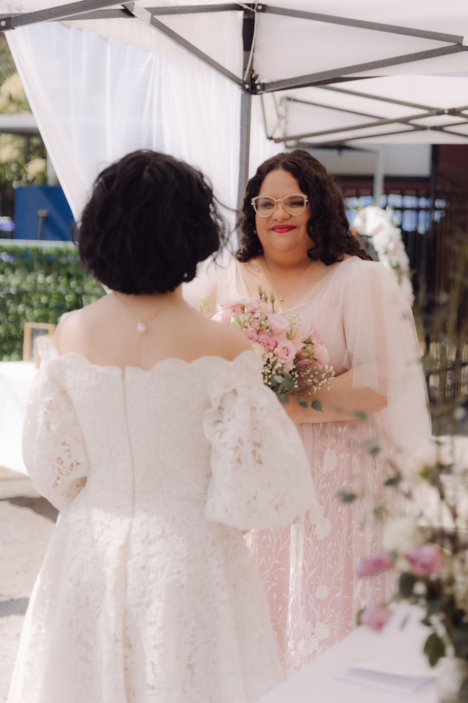 Two brides face each other at outdoor wedding ceremony, one in white lace, one in pink gown.