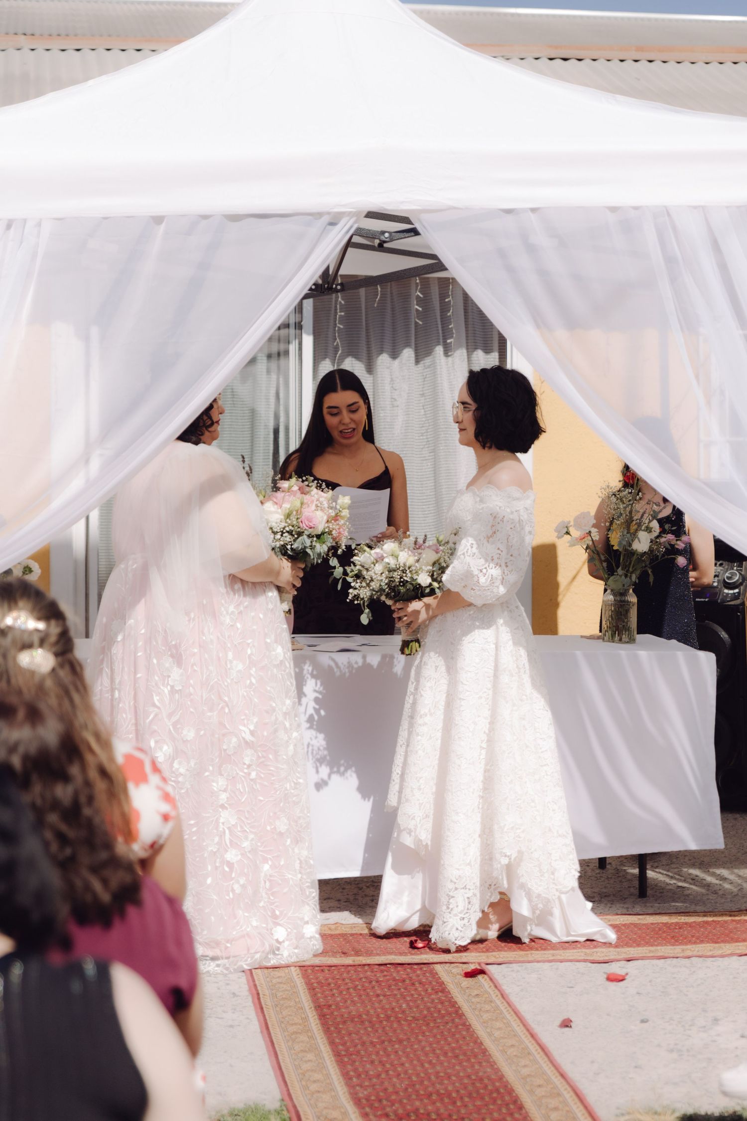 Two brides exchange vows under white canopy at outdoor wedding ceremony with officiant.