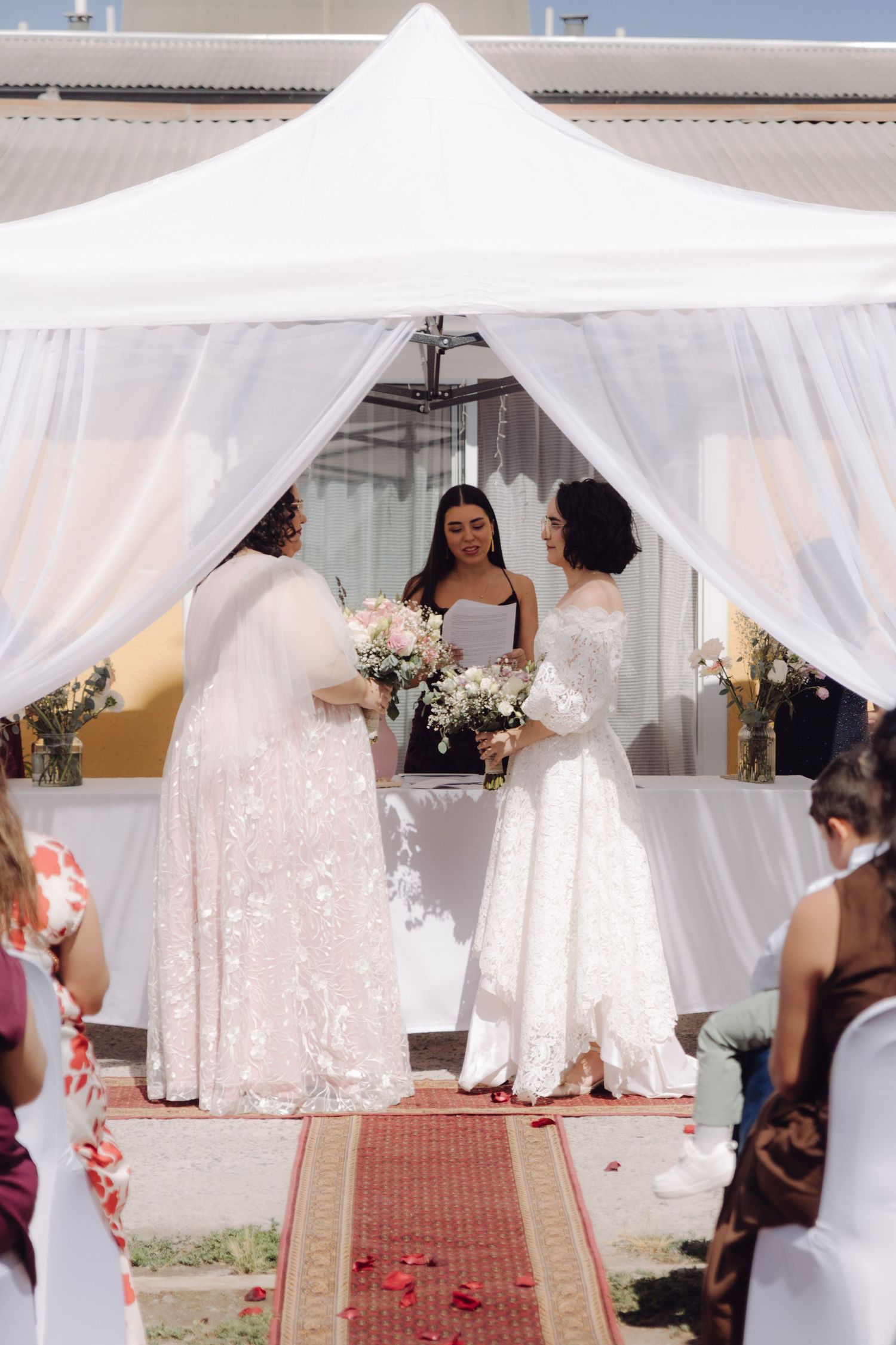Two brides stand at floral altar under white canopy during outdoor wedding ceremony.
