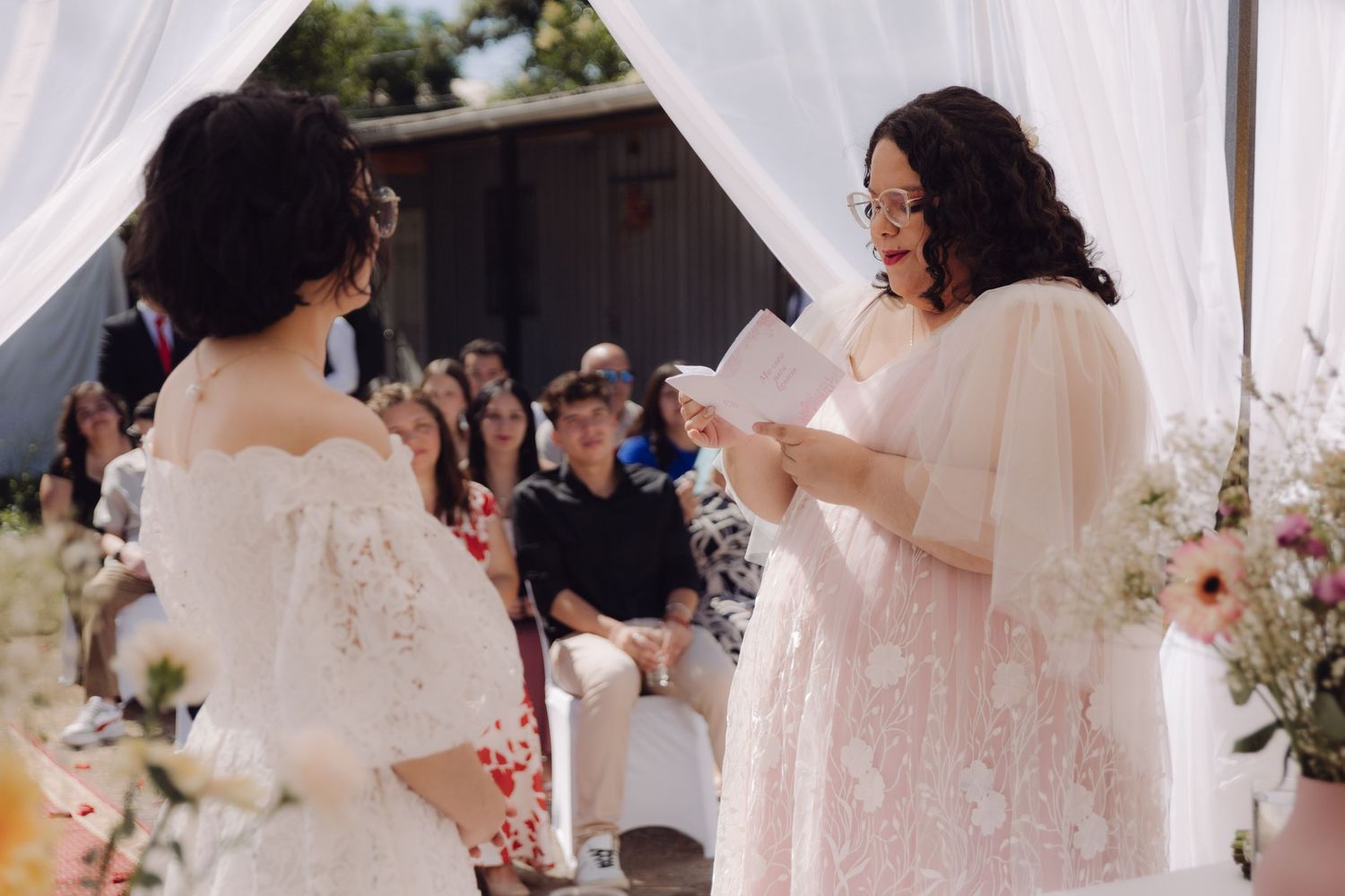 Bride in pink gown reads vows to partner in white lace dress at outdoor wedding ceremony.