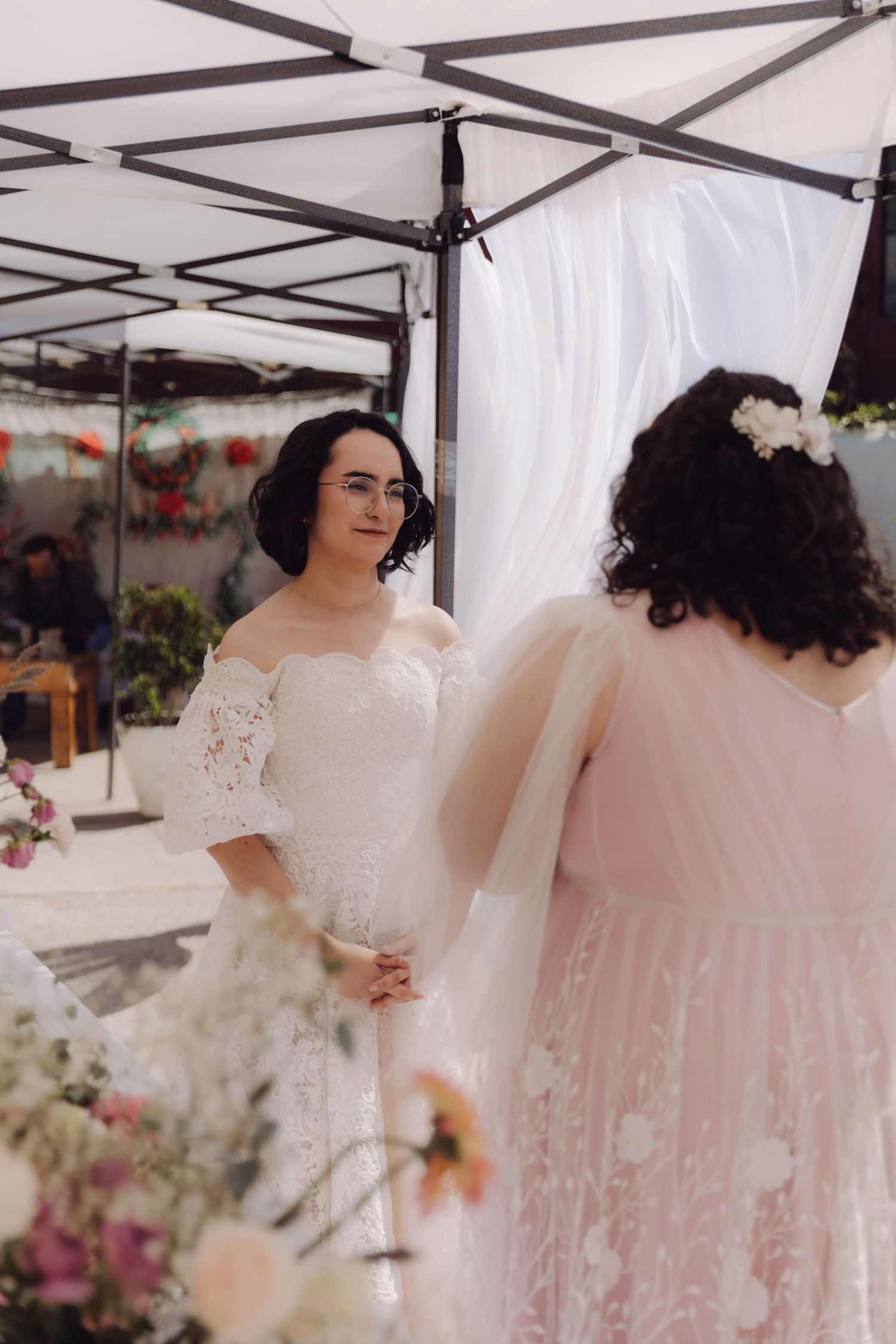 Two brides exchange vows at an outdoor wedding ceremony under a white canopy with floral decorations.