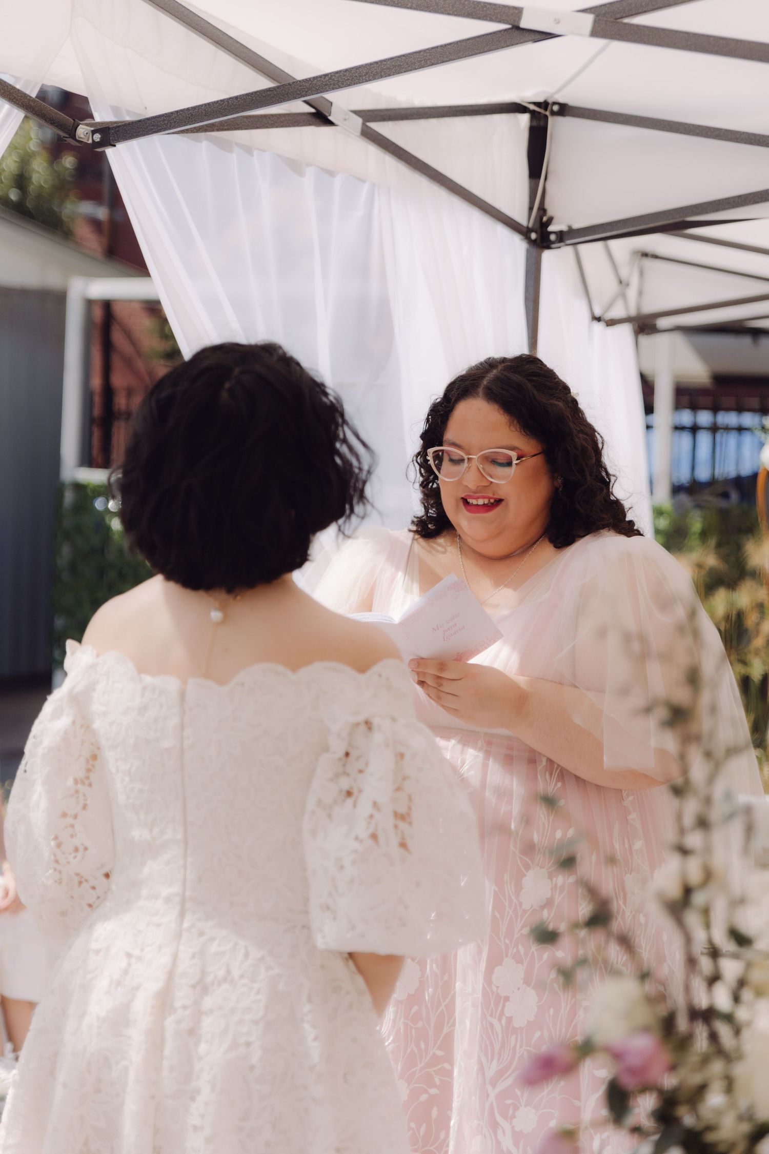Two brides share a joyful moment as one reads vows during their outdoor wedding ceremony.