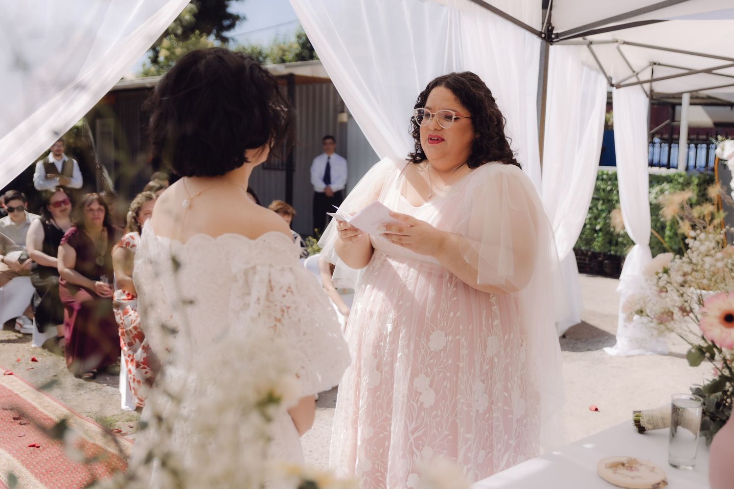 Bride in pink dress reads vows to her partner at an outdoor wedding with seated guests.