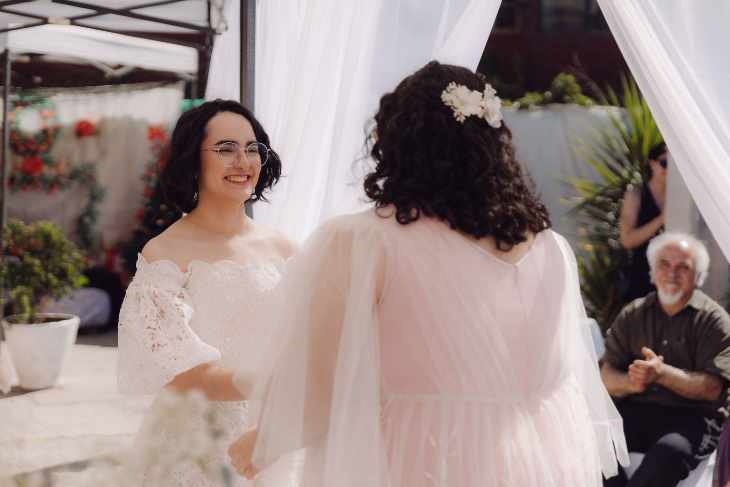 Two brides smile and hold hands during their outdoor wedding ceremony with guests in background.
