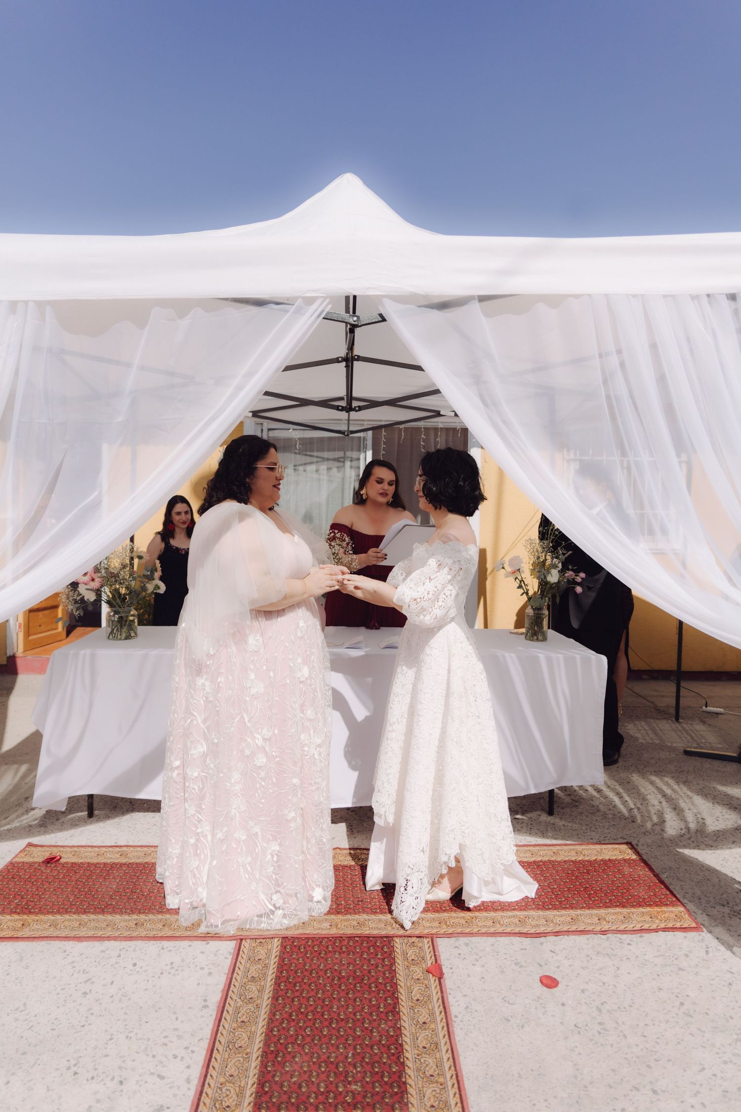 Two brides exchange rings under a white draped canopy at an outdoor wedding ceremony.