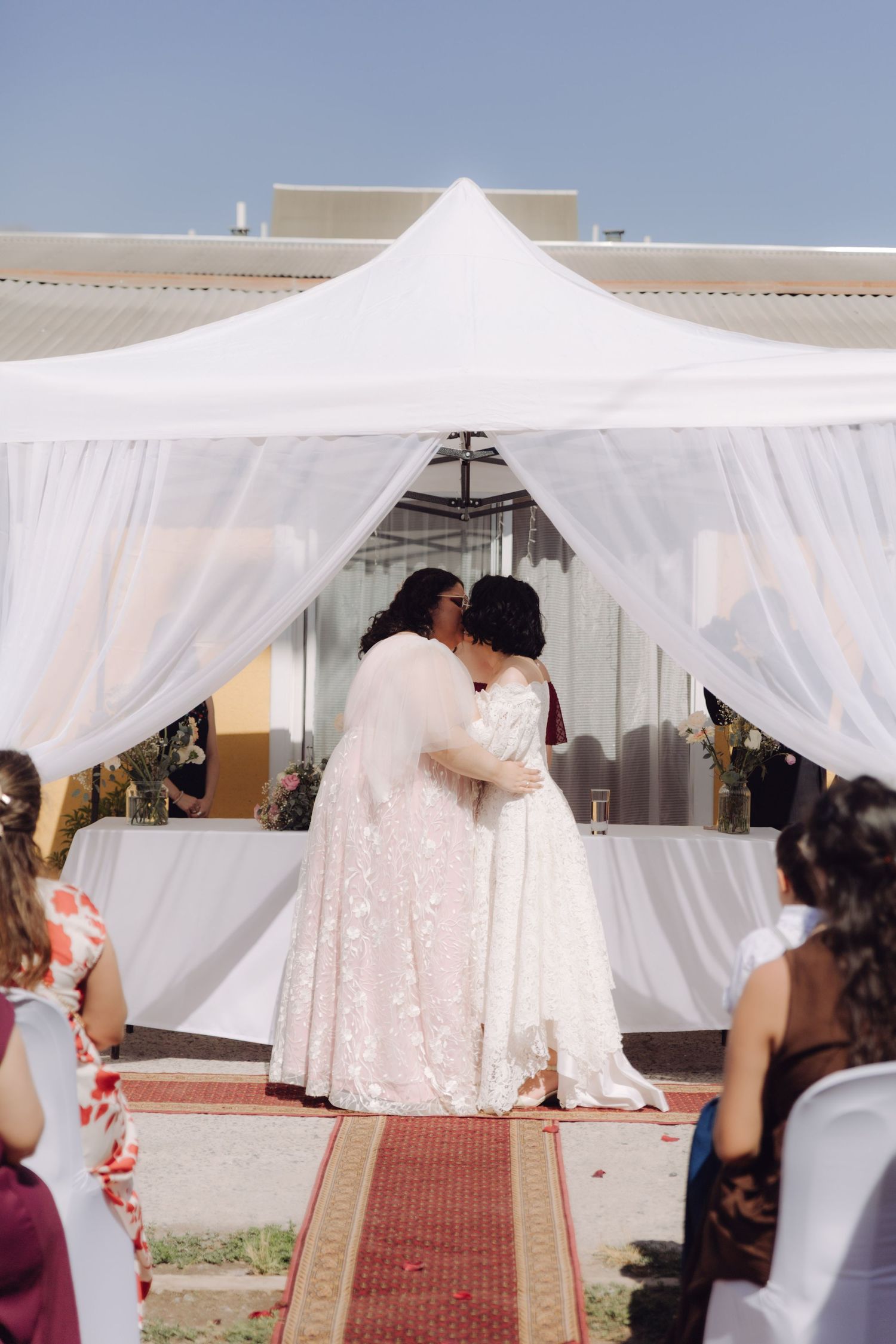 Two brides share their first kiss under a white canopy at their outdoor wedding ceremony.