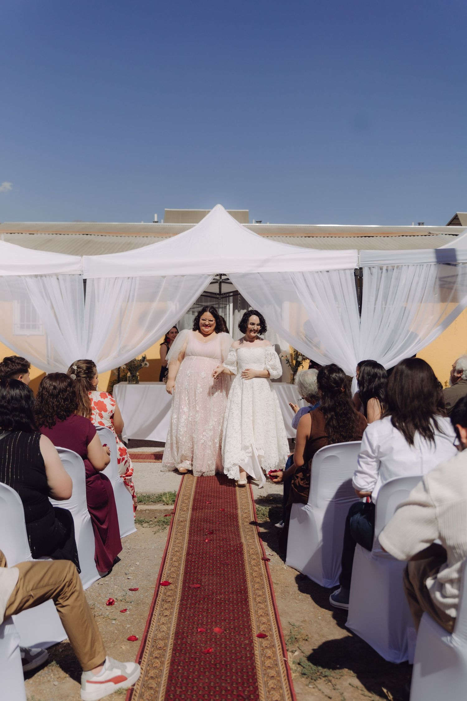 Two brides walk down outdoor wedding aisle in white lace gowns on red carpet under white canopy.