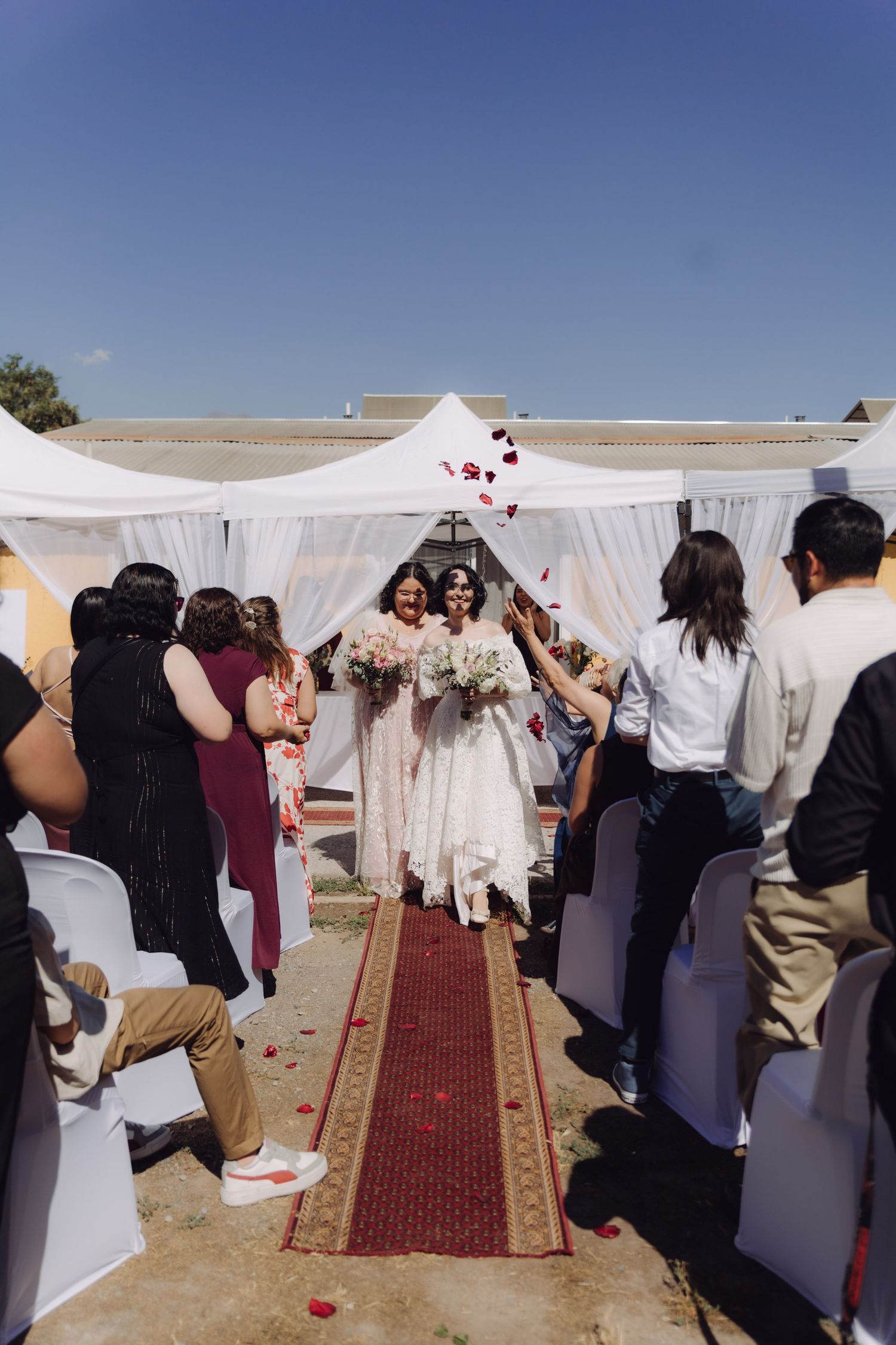 Guests toss rose petals as two brides walk down outdoor wedding aisle holding bouquets.