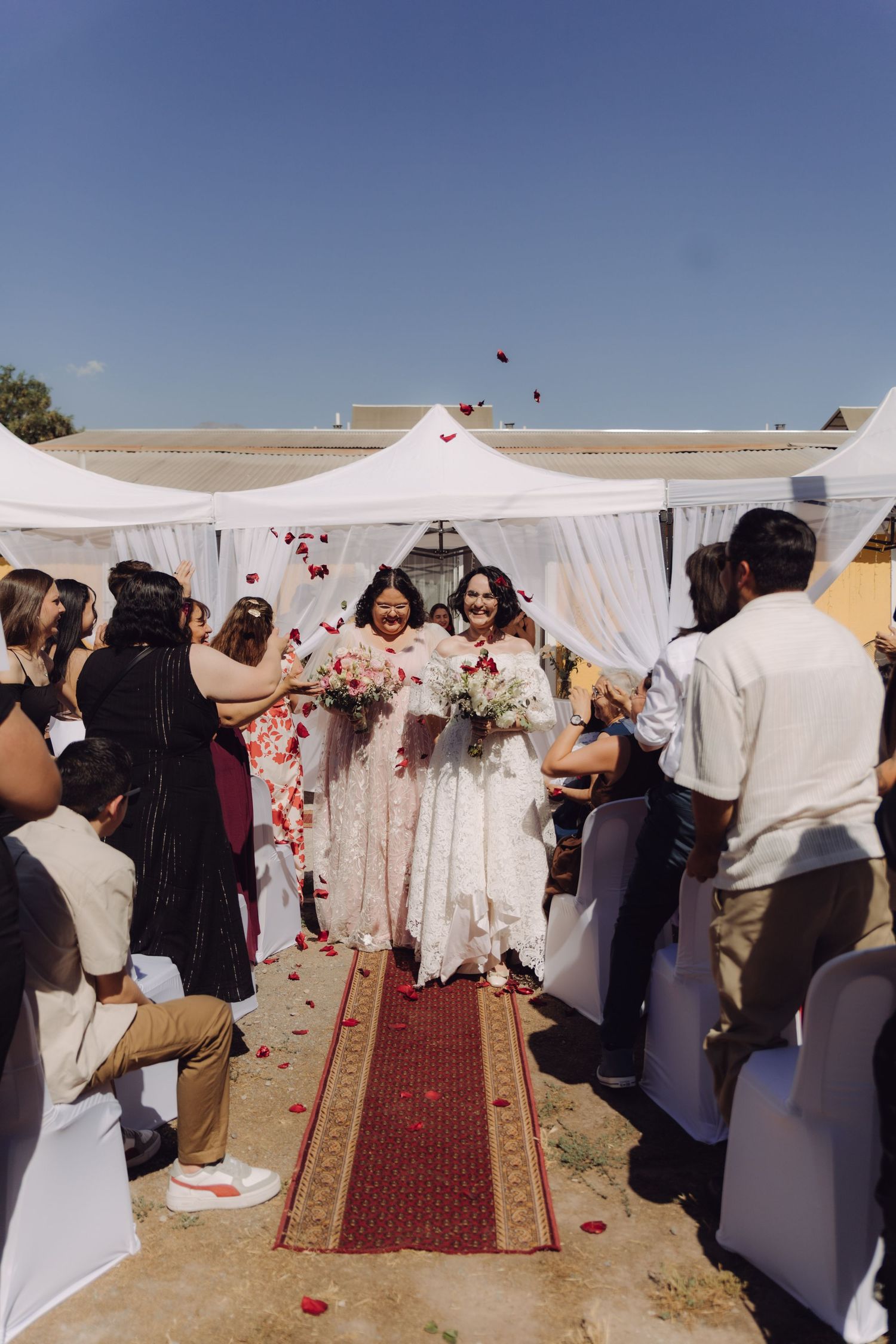 Smiling brides walk through shower of rose petals tossed by guests at outdoor wedding ceremony.