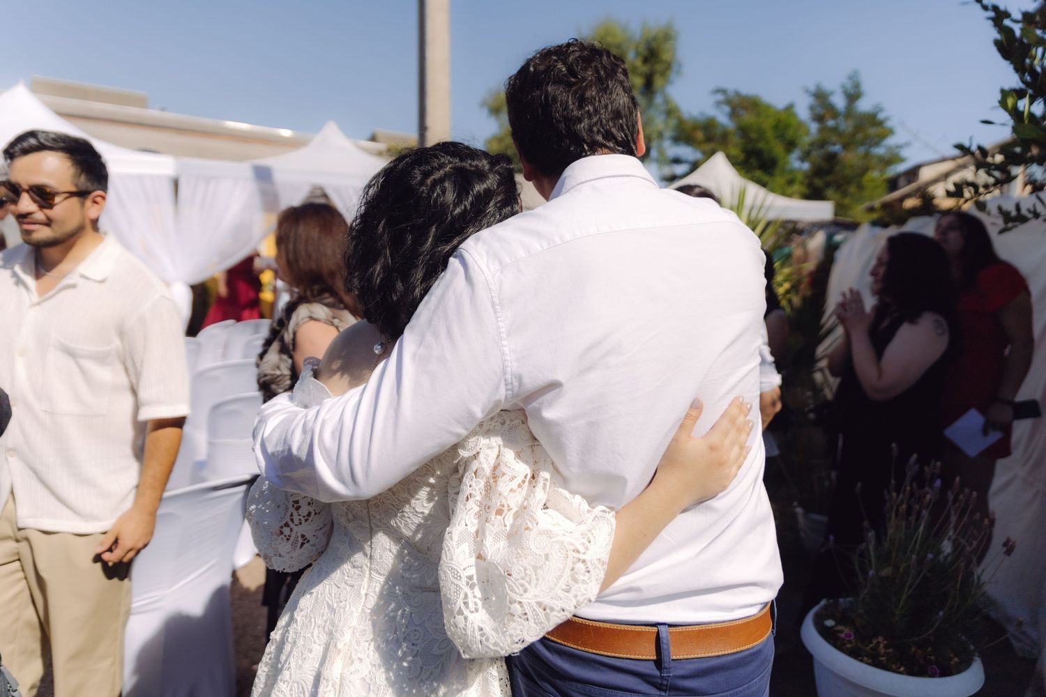 Wedding guest in white shirt embraces bride in lace gown at outdoor wedding reception.