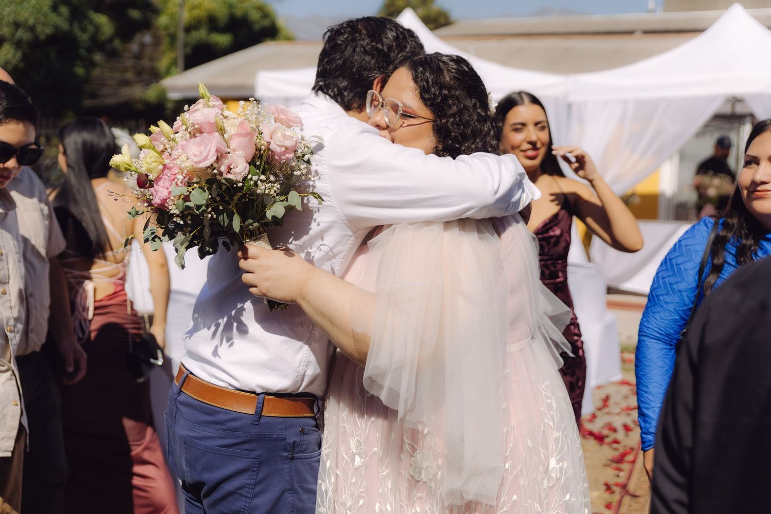 Bride holding pink floral bouquet shares warm embrace with guest in white shirt at wedding.