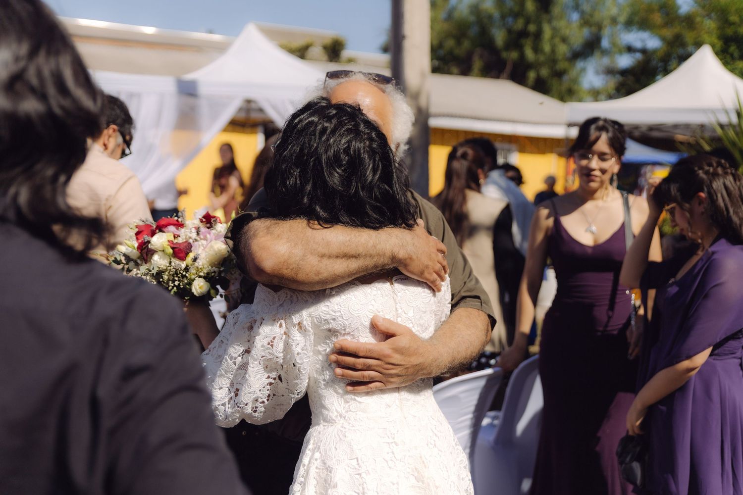 Bride in white lace gown embraces older male guest warmly at outdoor wedding celebration.