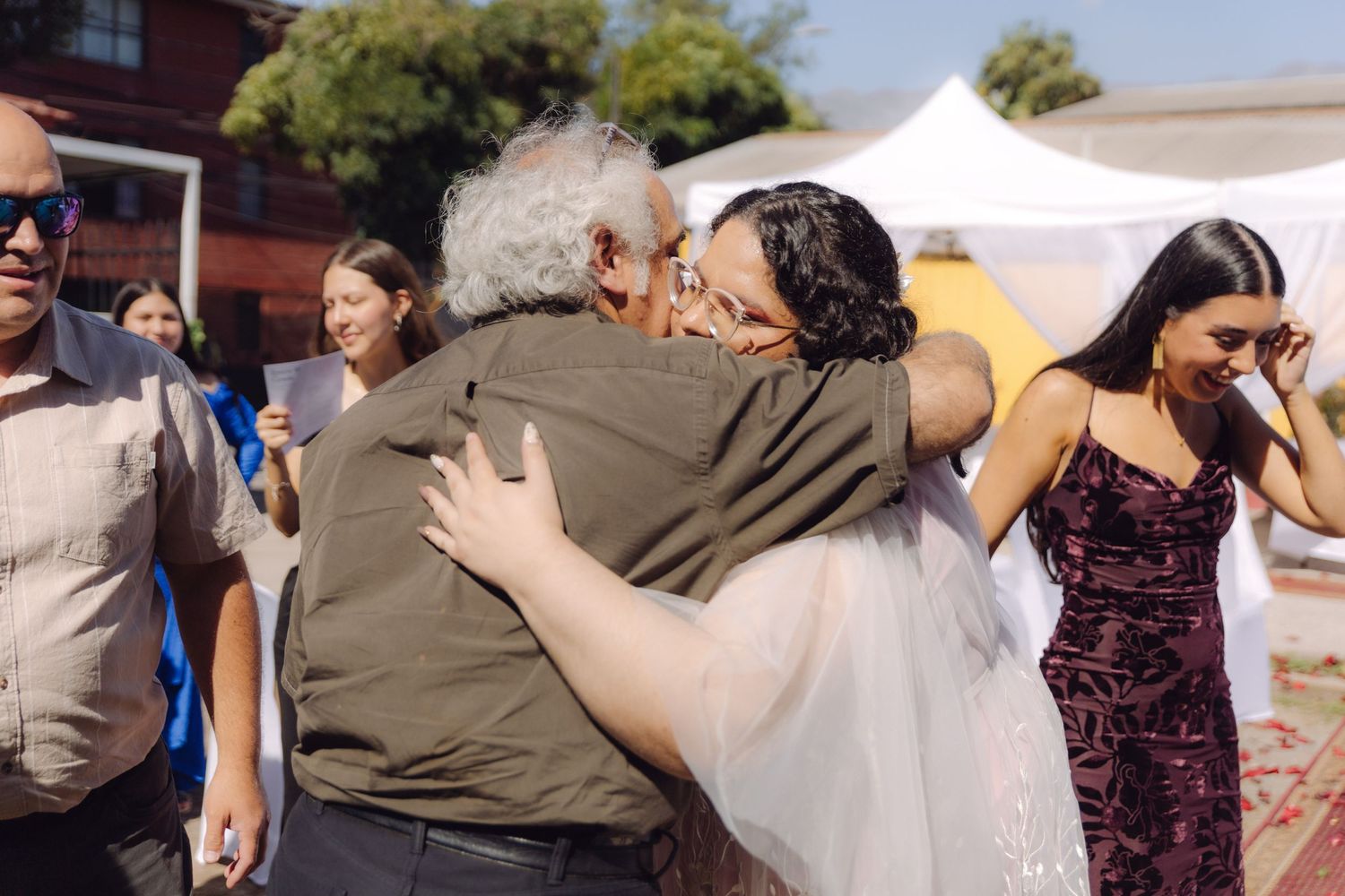 Bride hugs elderly guest at outdoor wedding while bridesmaids in purple dresses look on.