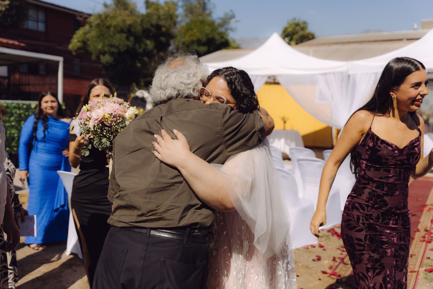 Bride in blush wedding gown embraces a guest warmly at an outdoor wedding ceremony.