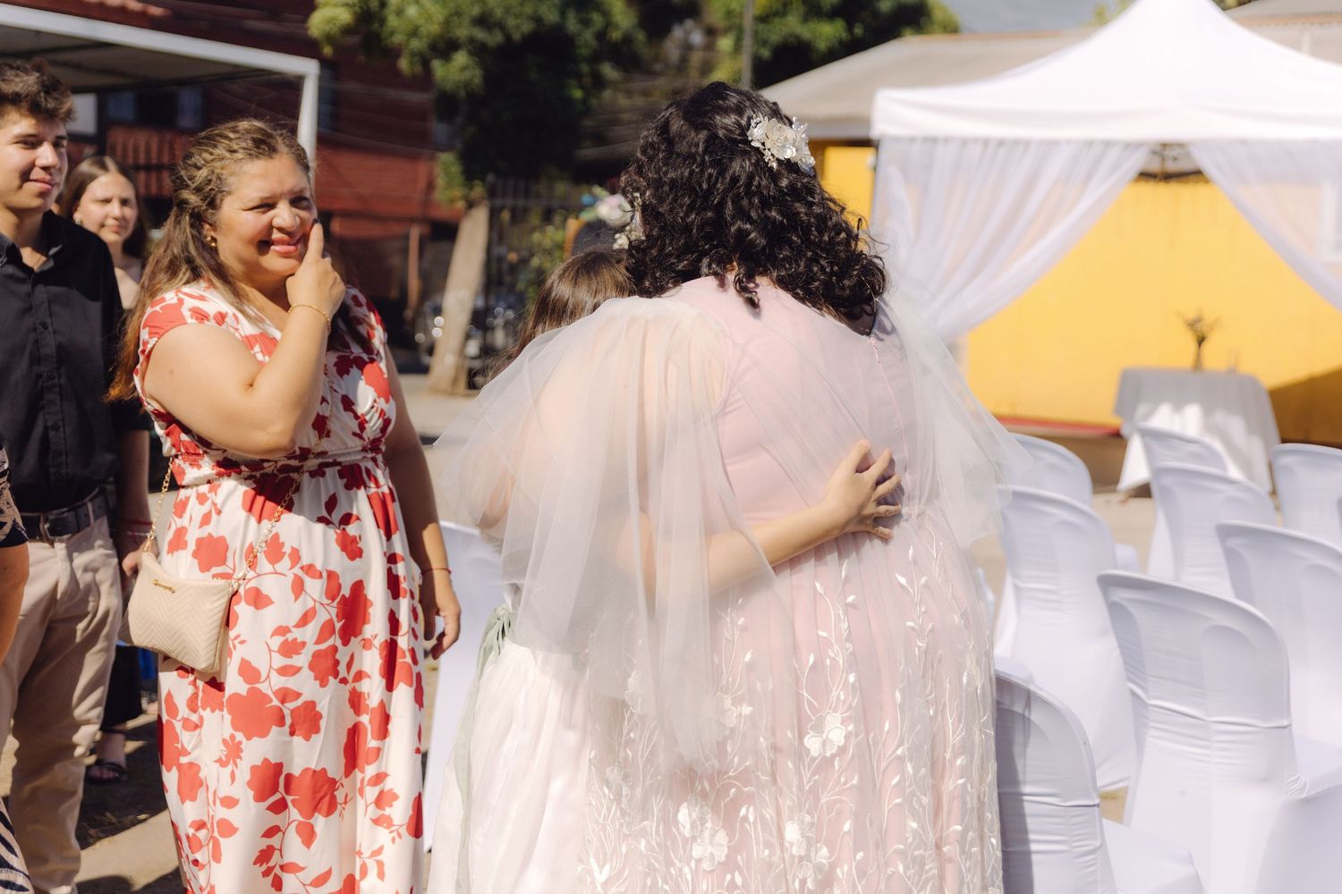 Emotional guest in floral dress embraces a veiled bride at an outdoor wedding celebration.