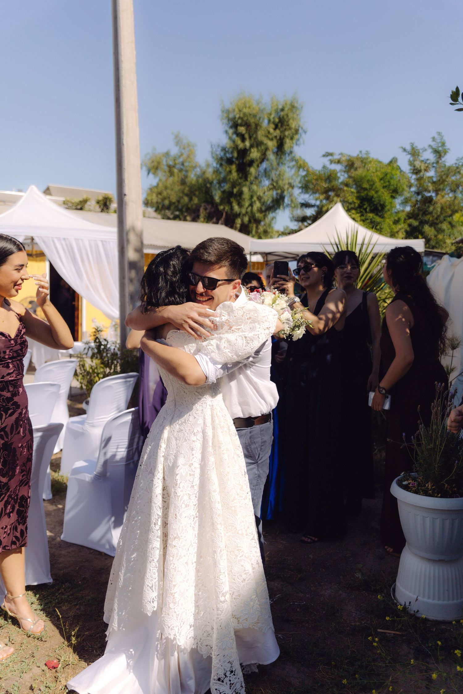 Bride in white lace gown hugs a guest warmly at an outdoor wedding ceremony with guests nearby.