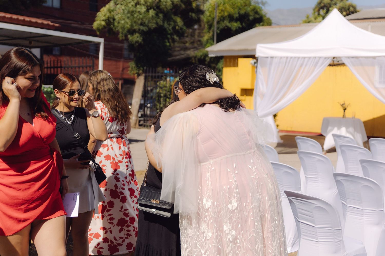 Bride in white lace gown greeted by guests at outdoor wedding ceremony with white chairs and floral arch.