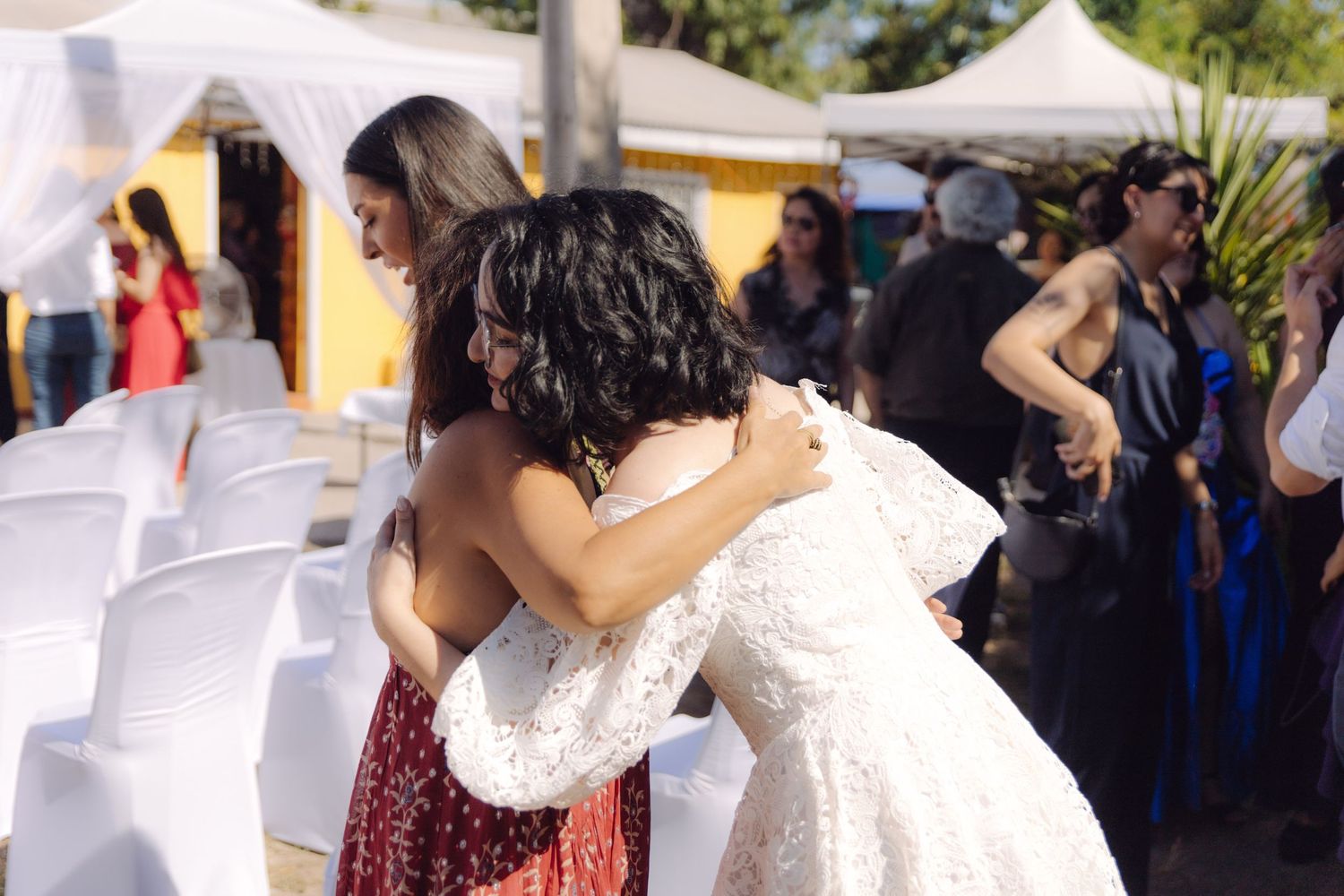 Bride in white lace wedding dress embracing a guest in burgundy dress at outdoor wedding reception.