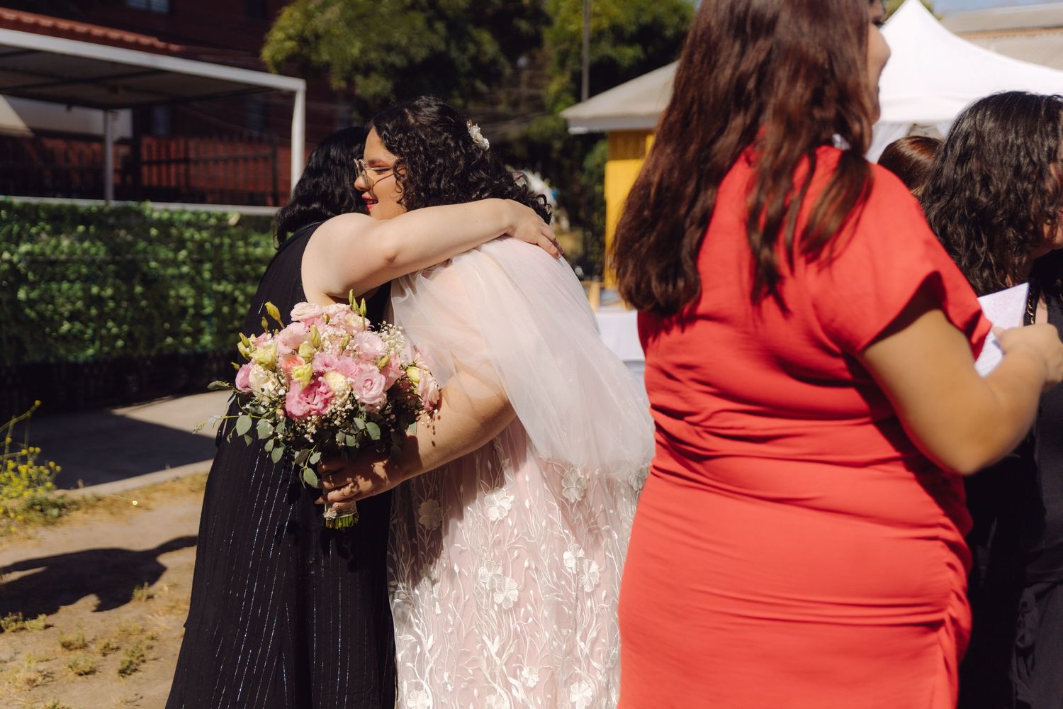 Bride holding pink bouquet receiving warm hug from dark-haired guest at sunny outdoor wedding.