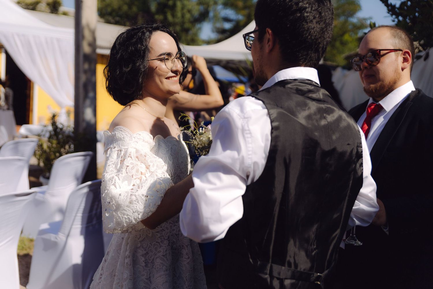Smiling bride in lace gown sharing joyful moment with groom in vest at outdoor wedding ceremony.