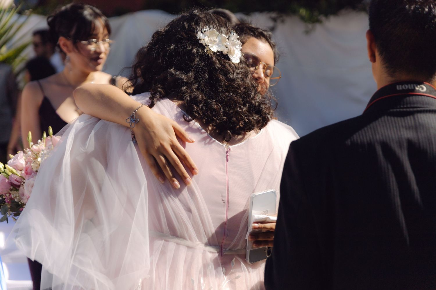 Bride in white tulle gown embracing guests while holding pink floral bouquet at outdoor wedding.