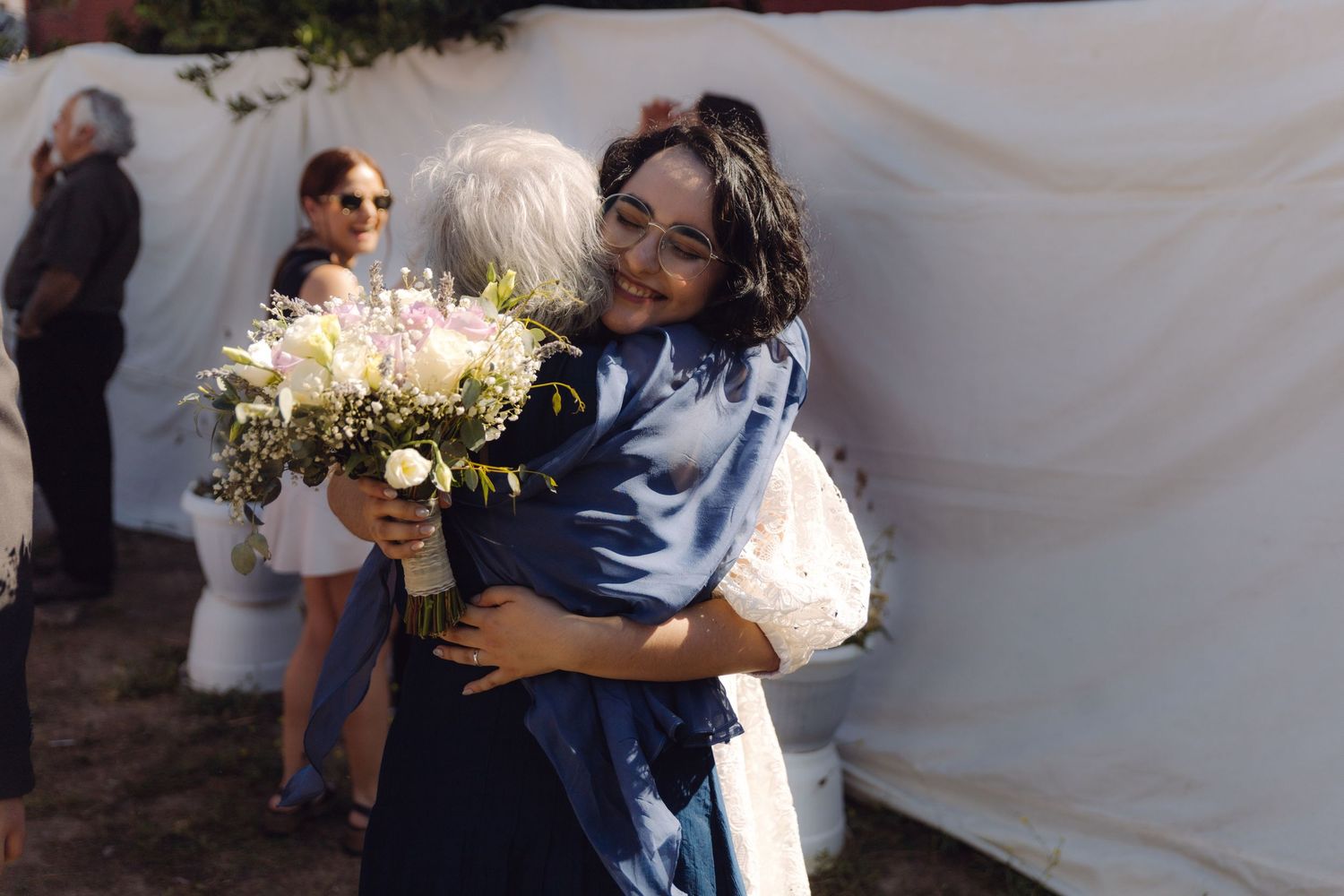 Bride in white lace dress hugging elderly guest in navy blue outfit at outdoor wedding reception.