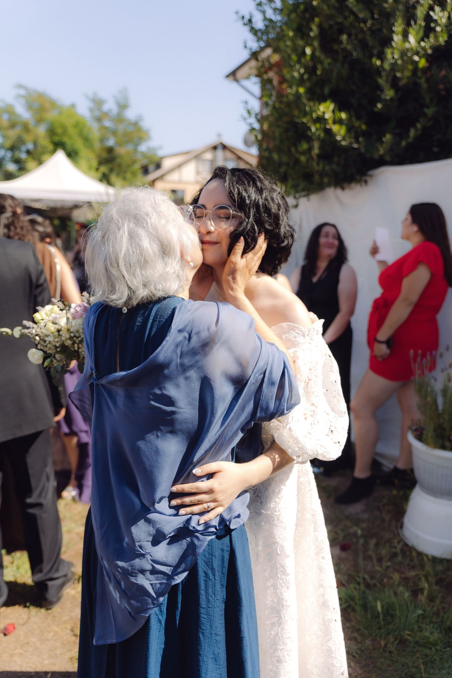 Elderly woman in blue dress kissing bride in white lace gown on cheek at outdoor wedding.