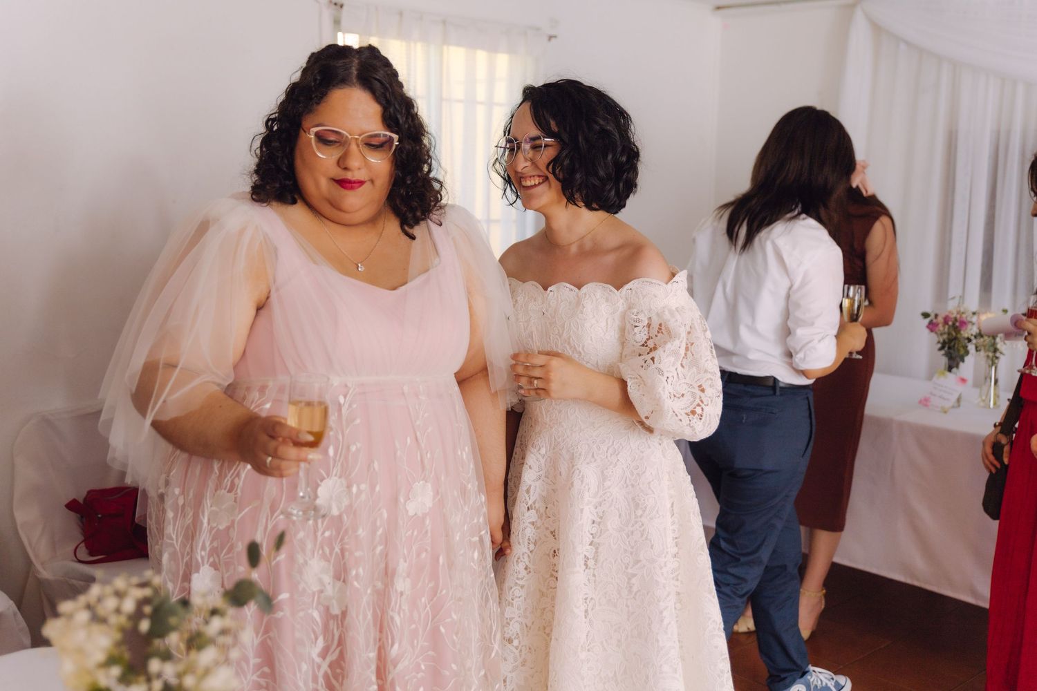 Two women in pink and white lace gowns holding champagne glasses together at wedding reception.