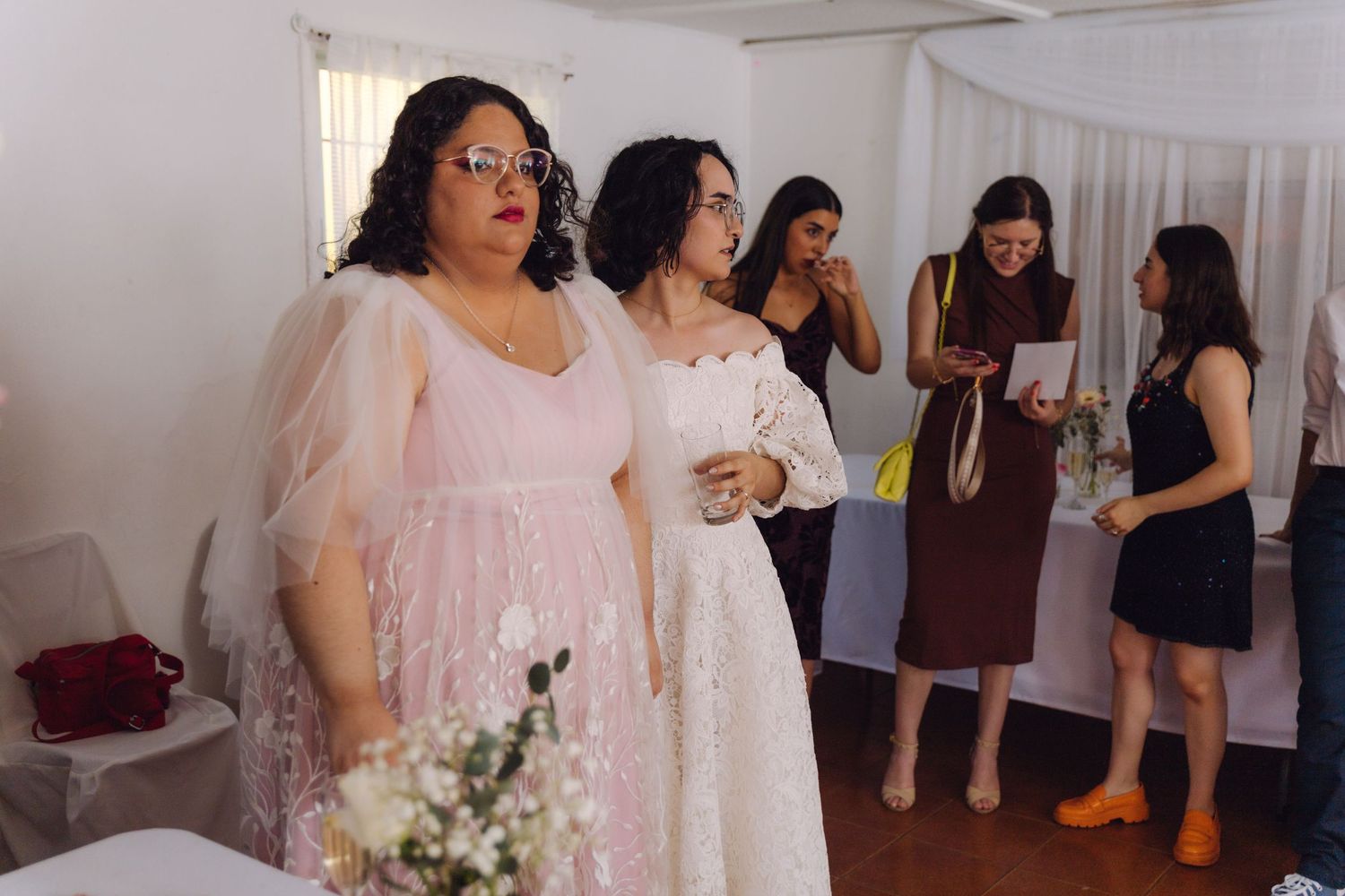 Two elegantly dressed women in pink and white gowns socializing at indoor wedding reception.