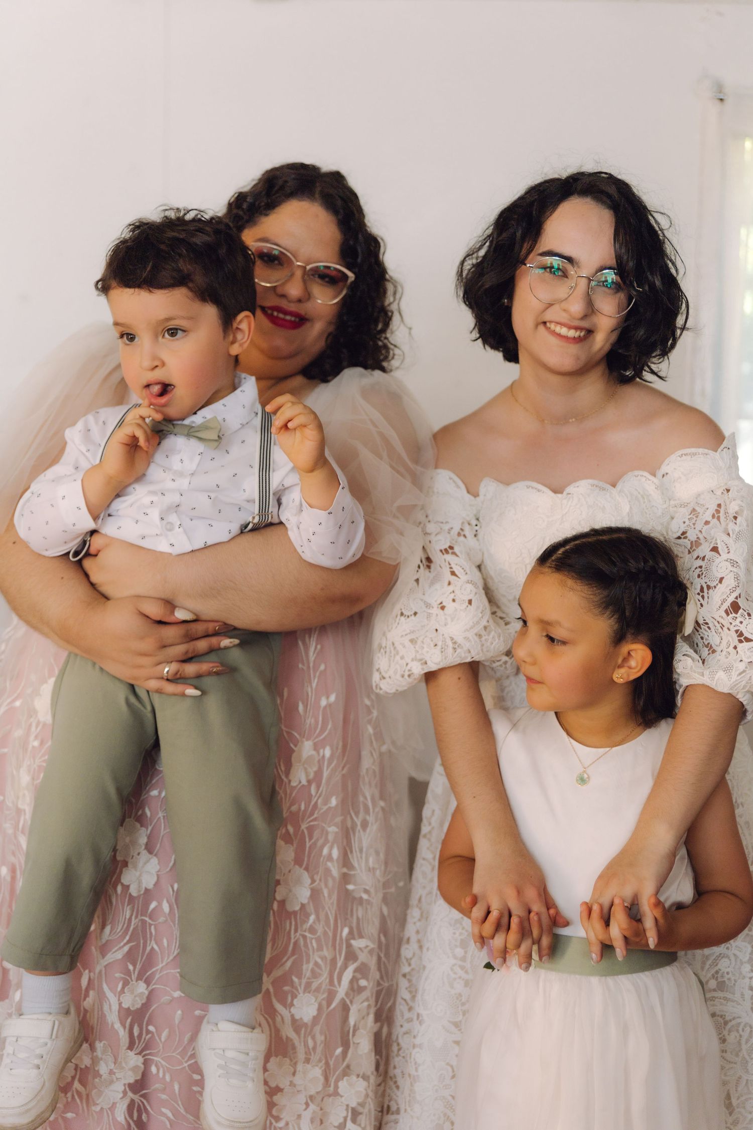 Two brides in pink and white lace gowns posing with young boy and girl at wedding celebration.