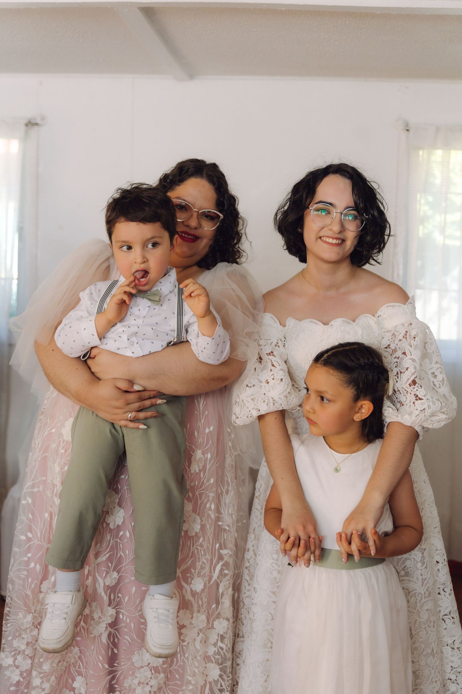 Two brides in white and pink lace gowns pose with their children at a wedding celebration.