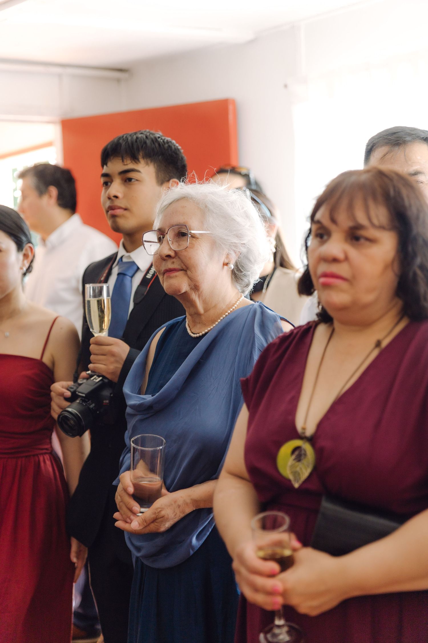 Elegantly dressed wedding guests holding champagne glasses watch a ceremony at an indoor venue.