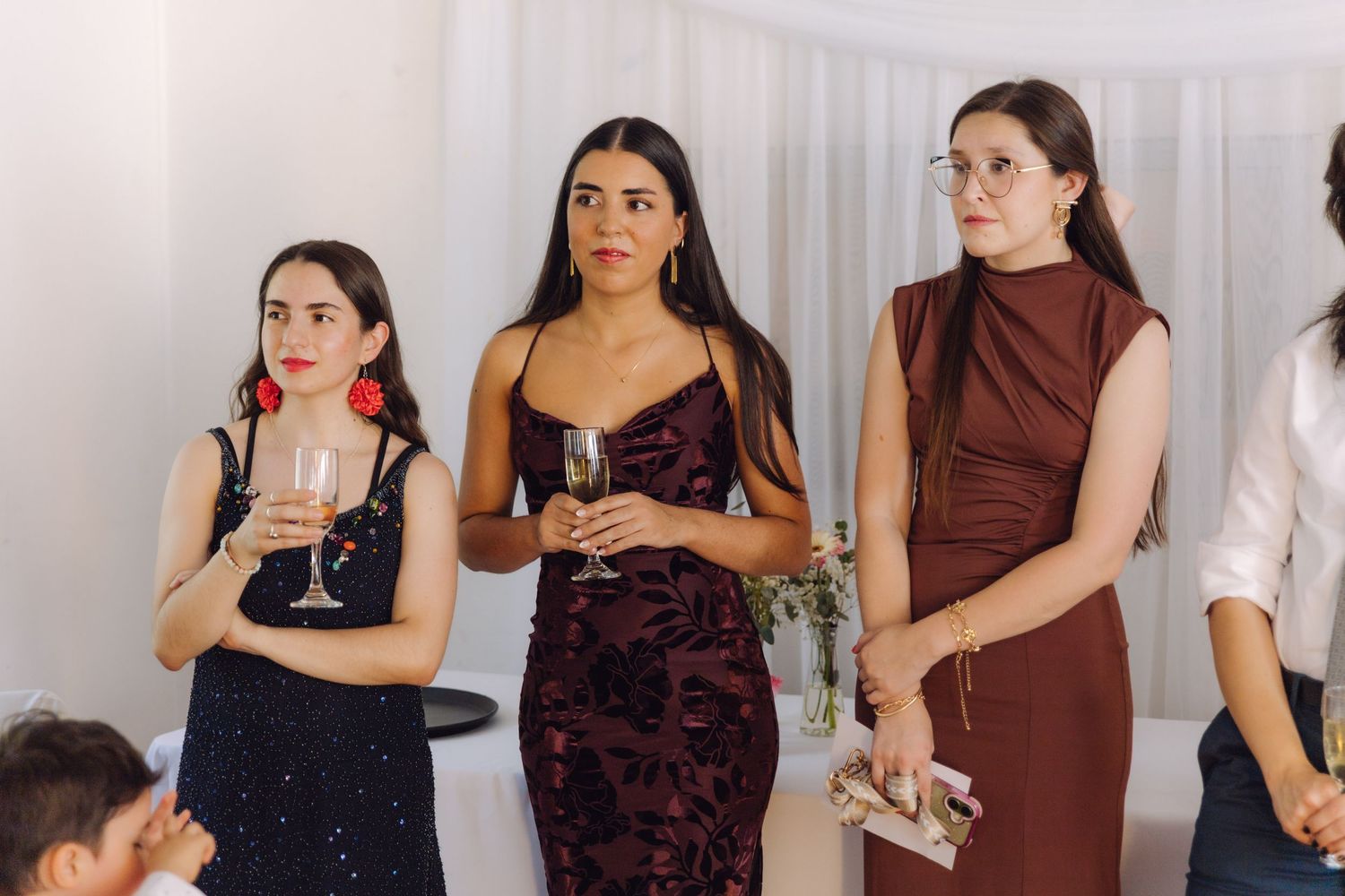 Three women in elegant dresses hold champagne glasses at a formal wedding reception event.