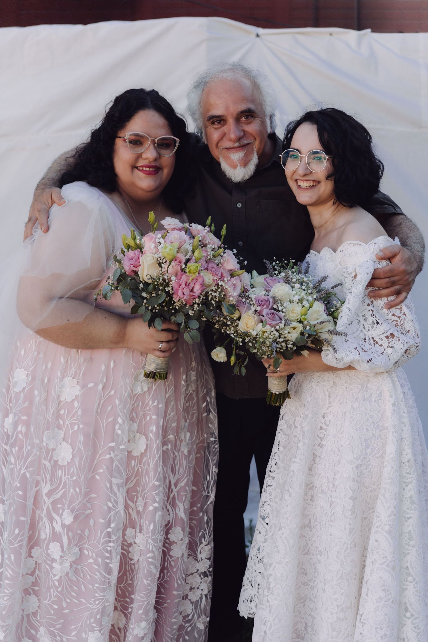 Two brides in lace gowns holding bouquets pose with an older man at their wedding.